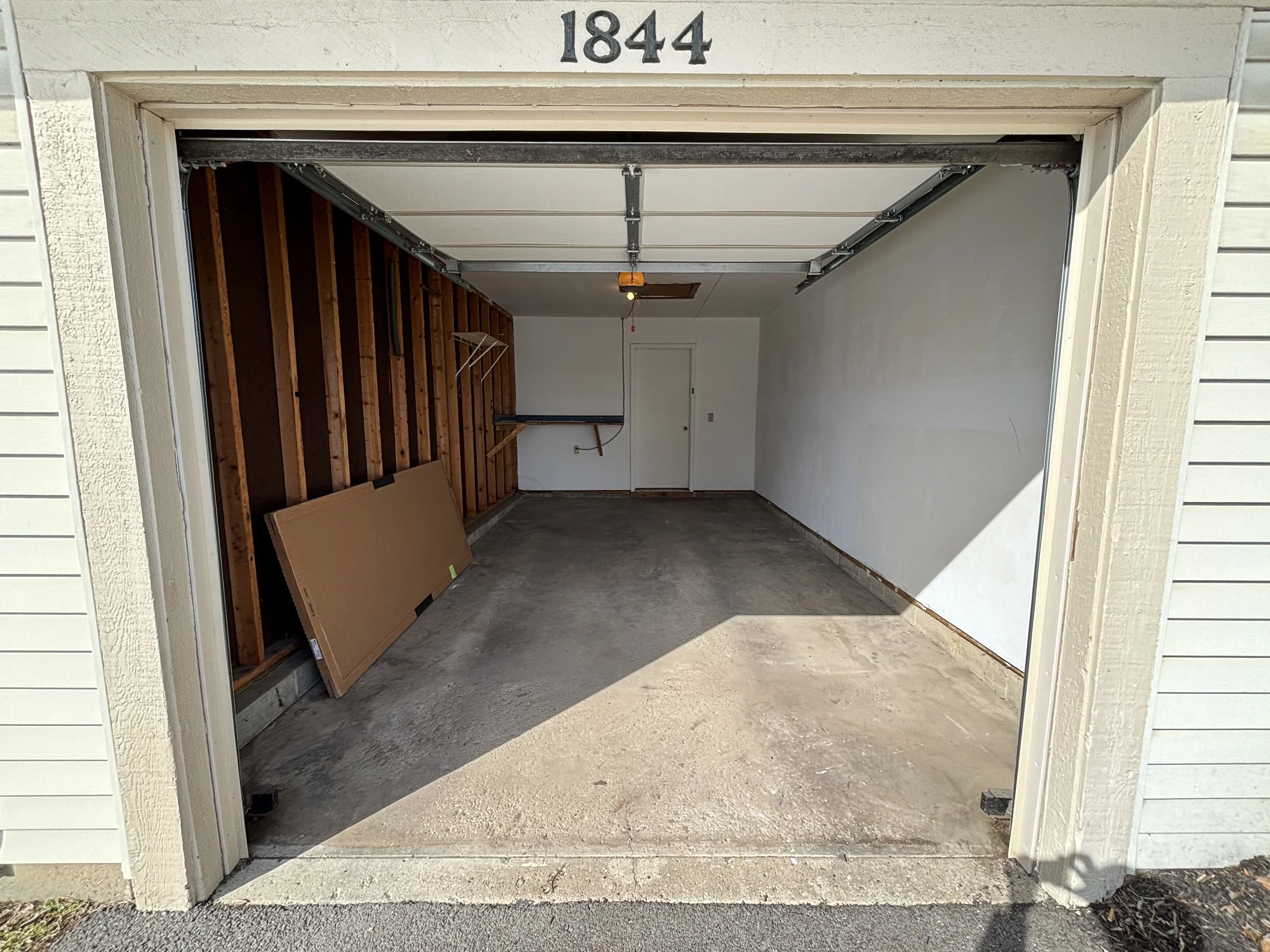 Empty single-car garage with open door, concrete floor, white walls, and a wooden panel on the left wall. The garage has a ceiling-mounted garage door opener and a small white door at the back.