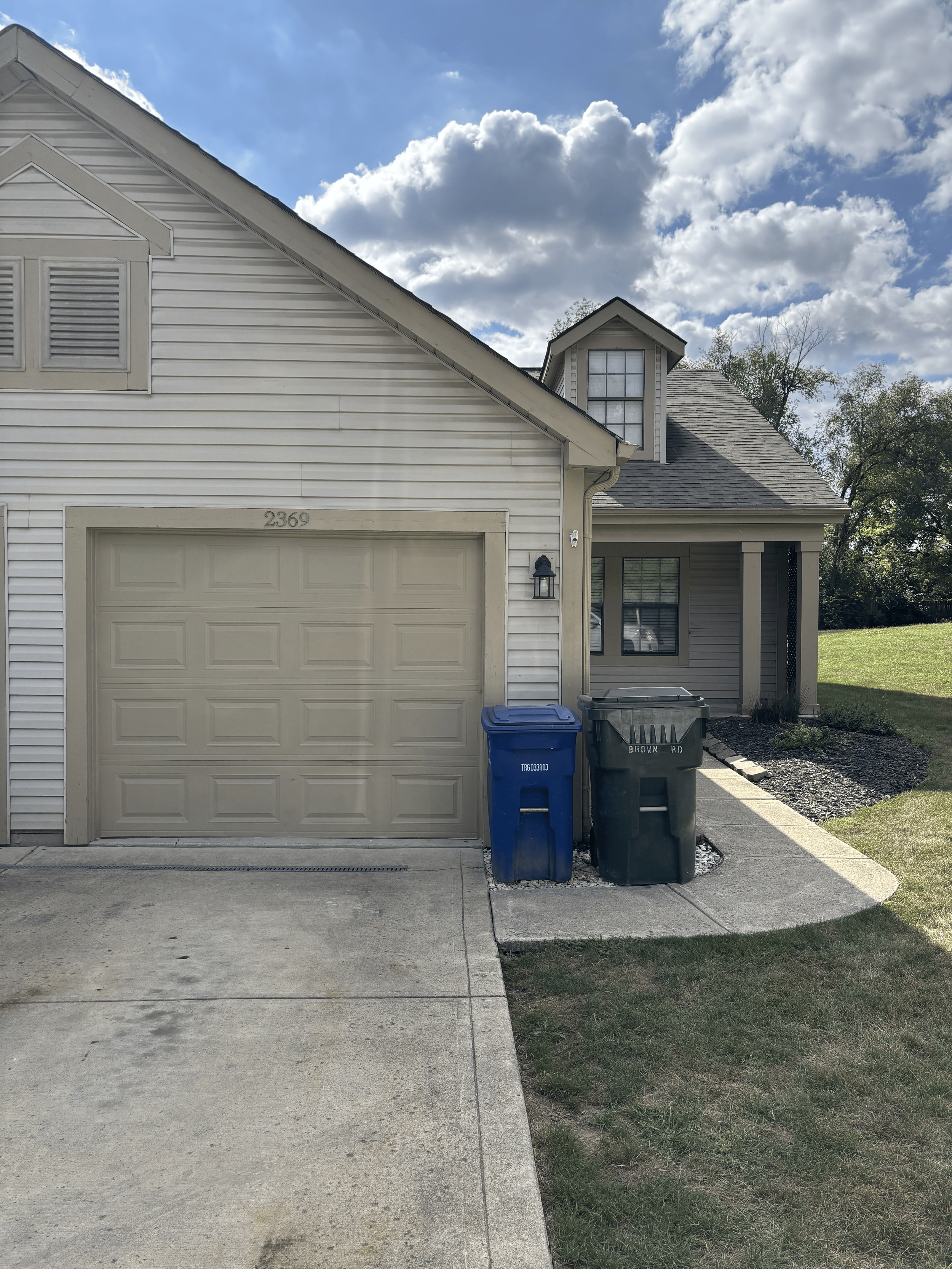 Front of a house with a garage, two trash bins, a porch, and a grassy yard. Blue sky with scattered clouds.