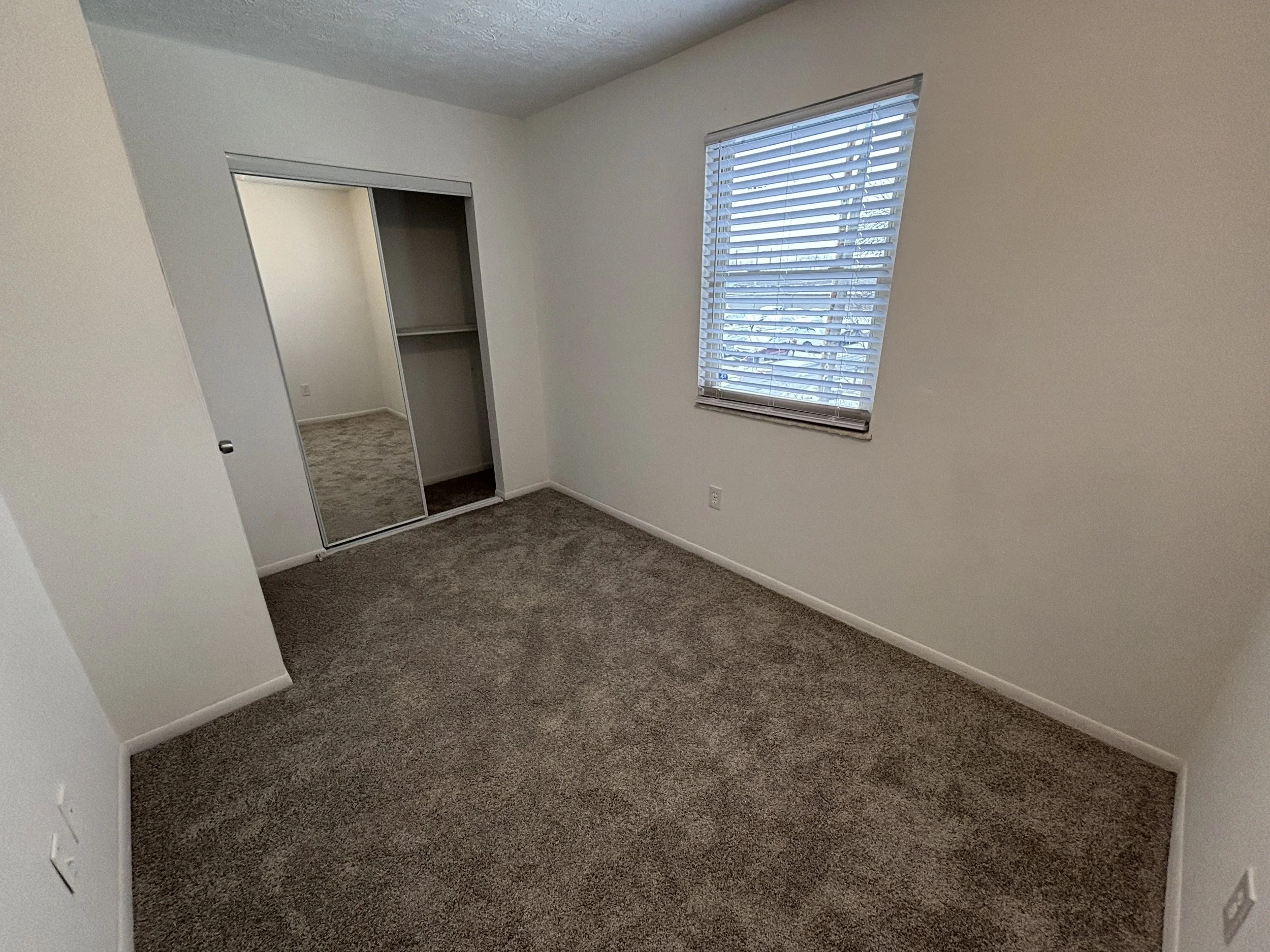 Empty bedroom with beige carpet, white walls, window with blinds, and a closet with sliding mirror doors.