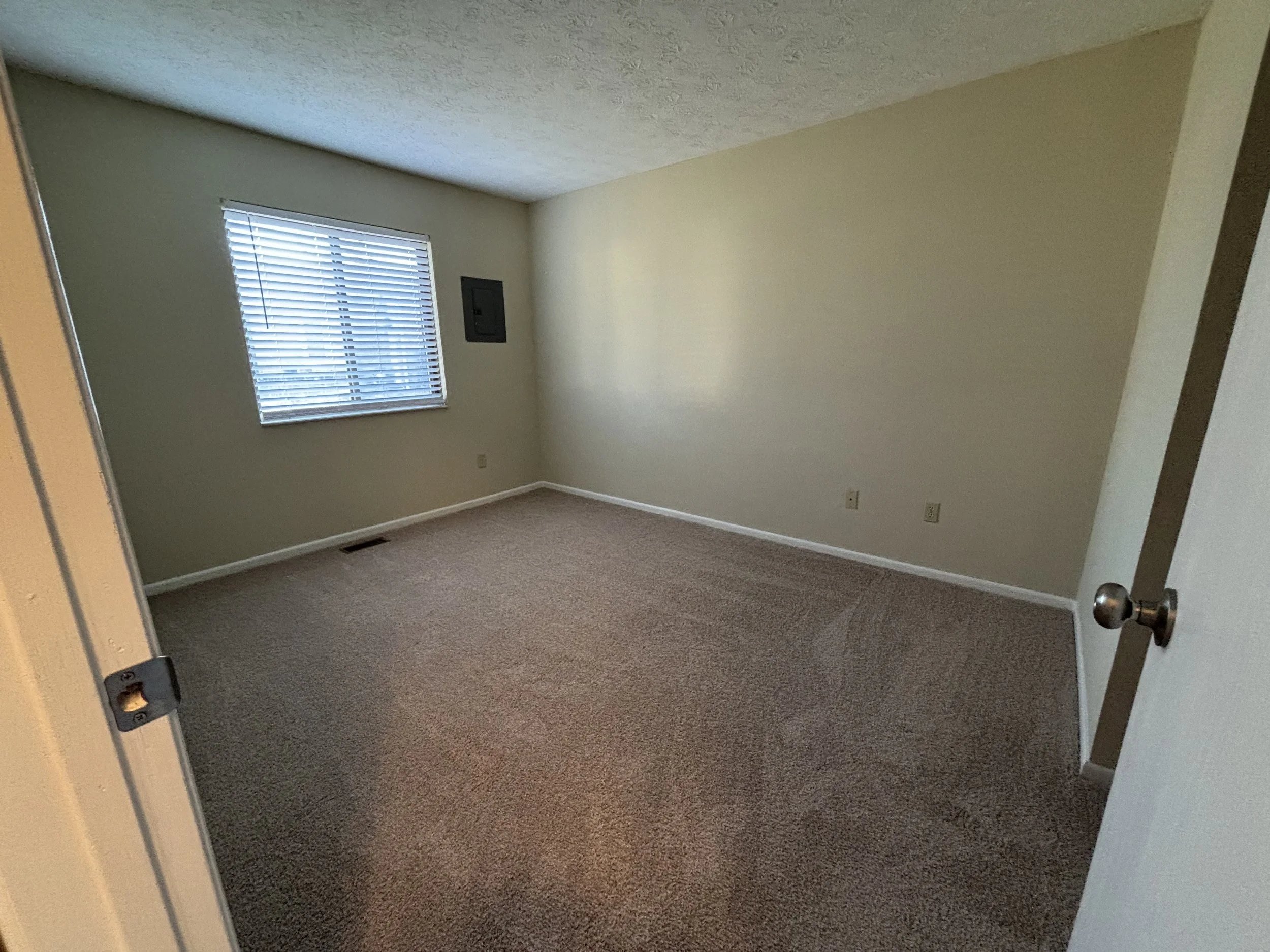 Empty room with beige carpet, off-white walls, a window with horizontal blinds, and a wall-mounted electrical panel.