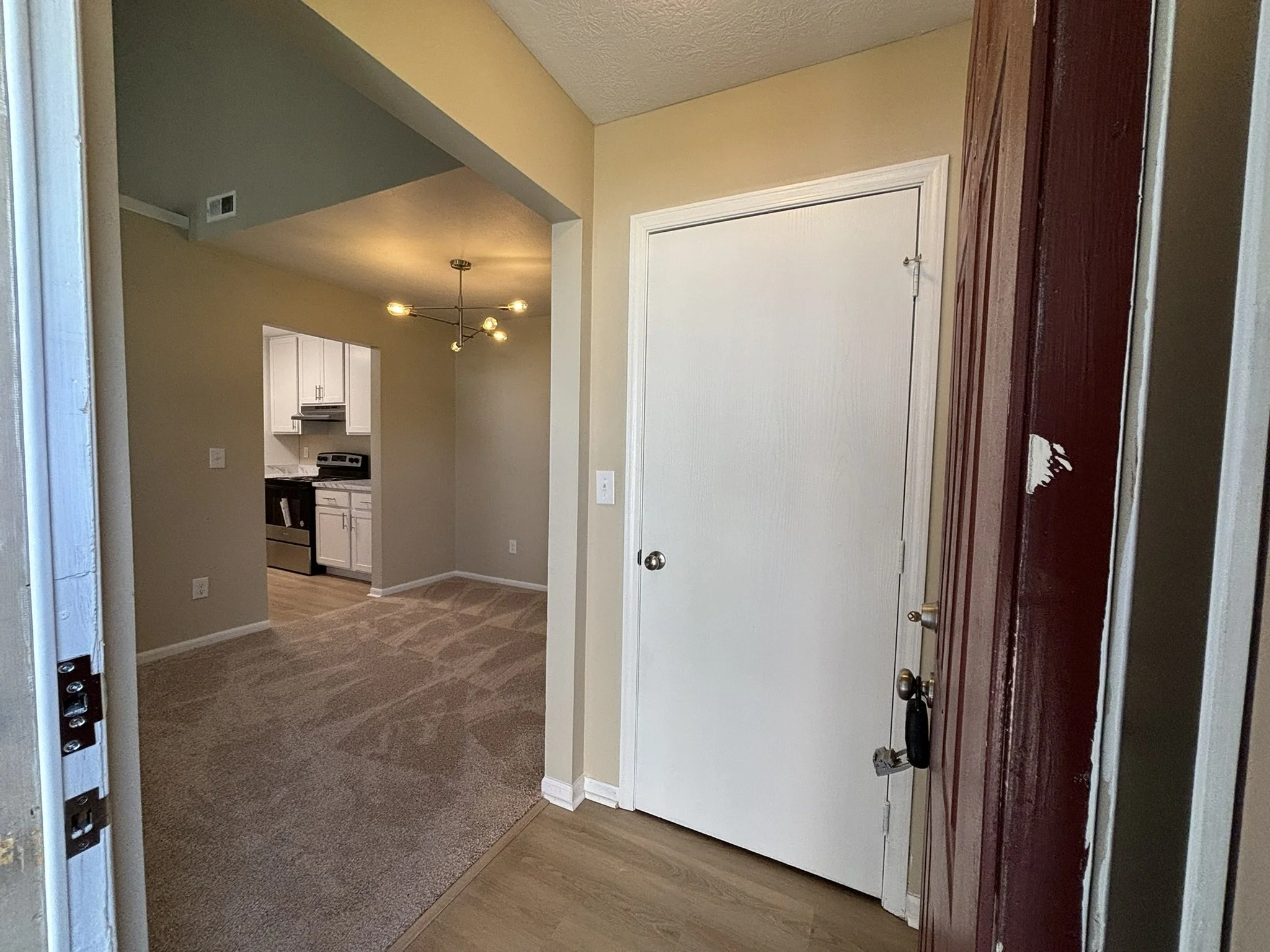 Inside view of a house entrance showing a doorway, a white door, a kitchen with white cabinets, a beige wall, a modern ceiling light fixture, and a carpeted floor.