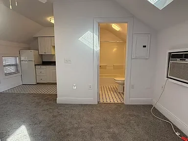 Living area with carpet, kitchen with white cabinets and appliances, and a bathroom with a toilet visible through an open door.