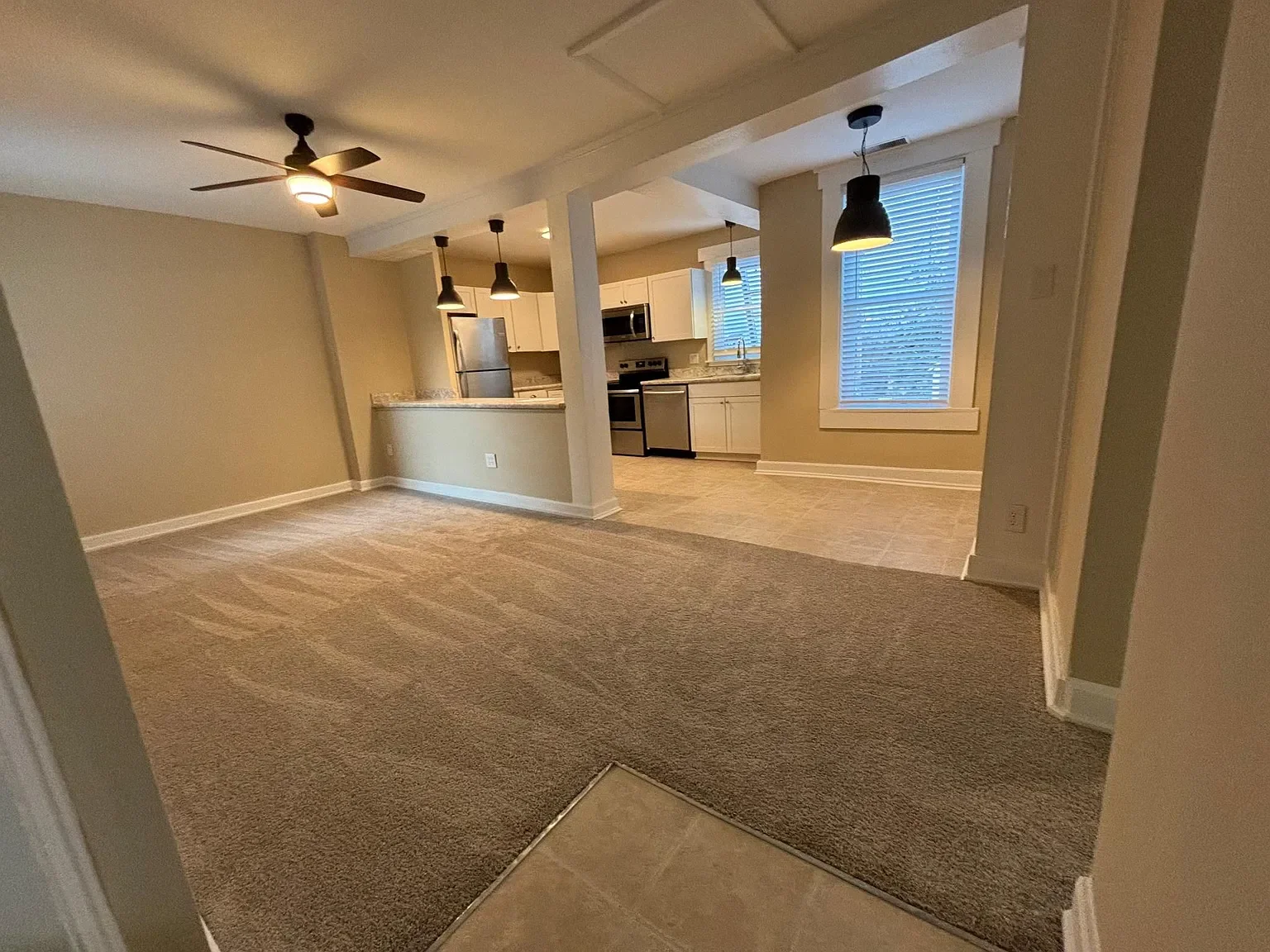 Interior view of an open floor plan living room and kitchen area with beige walls, gray carpeting, tiled flooring, modern light fixtures, and large windows with blinds.