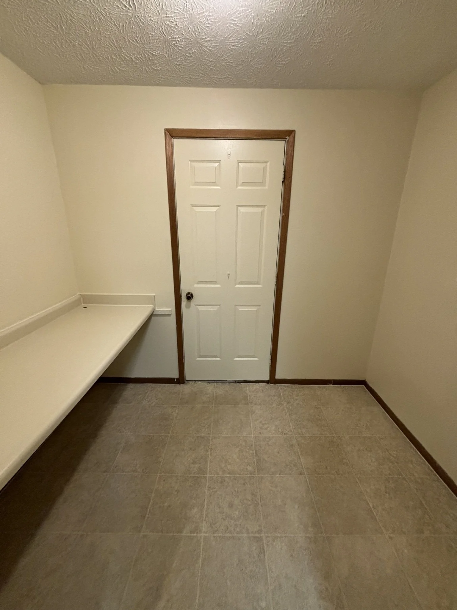 Empty room with beige tiled floor, white walls, a white door with wooden trim, and a built-in beige countertop on the left side.