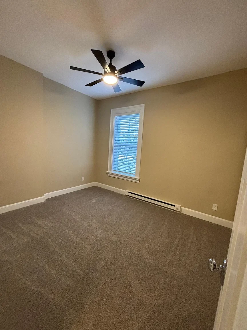 Empty bedroom with beige walls, gray carpet, white trim, a ceiling fan, and a window with blinds.