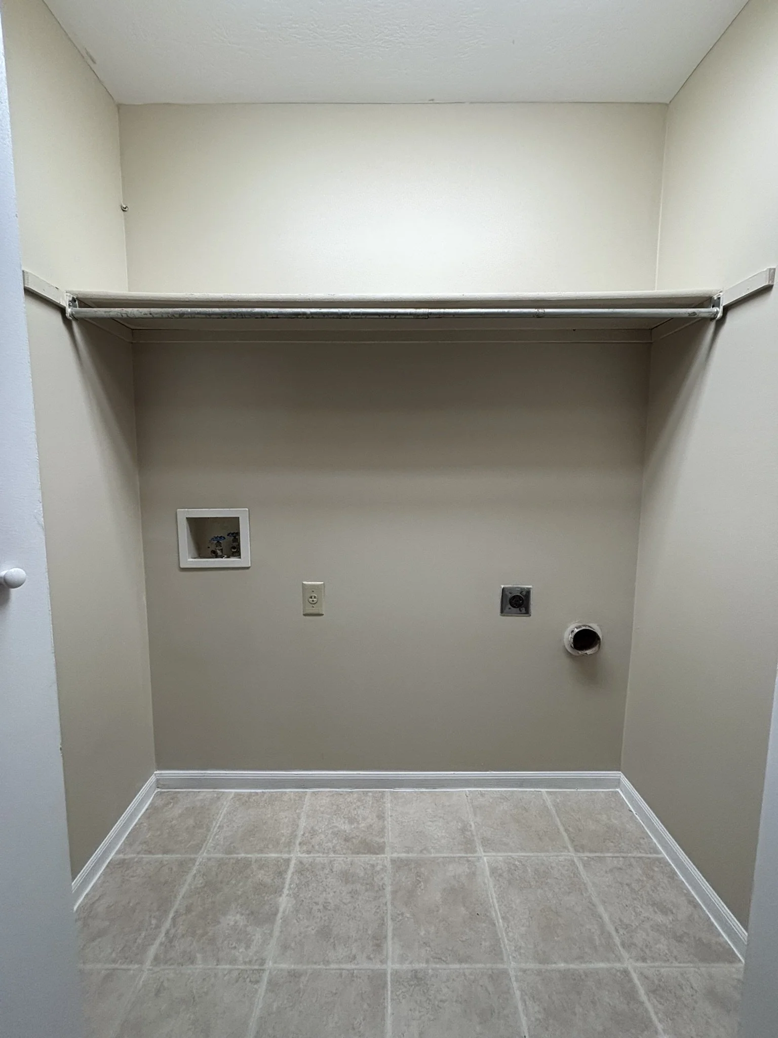 Empty laundry room with beige walls, tiled floor, a metal shelf, electrical outlets, a water supply box, and a vent pipe.