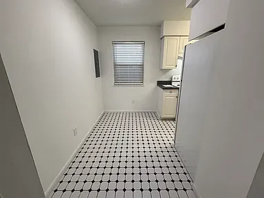 Empty kitchen with white appliances, a window with blinds, white cabinets, and a patterned tile floor.