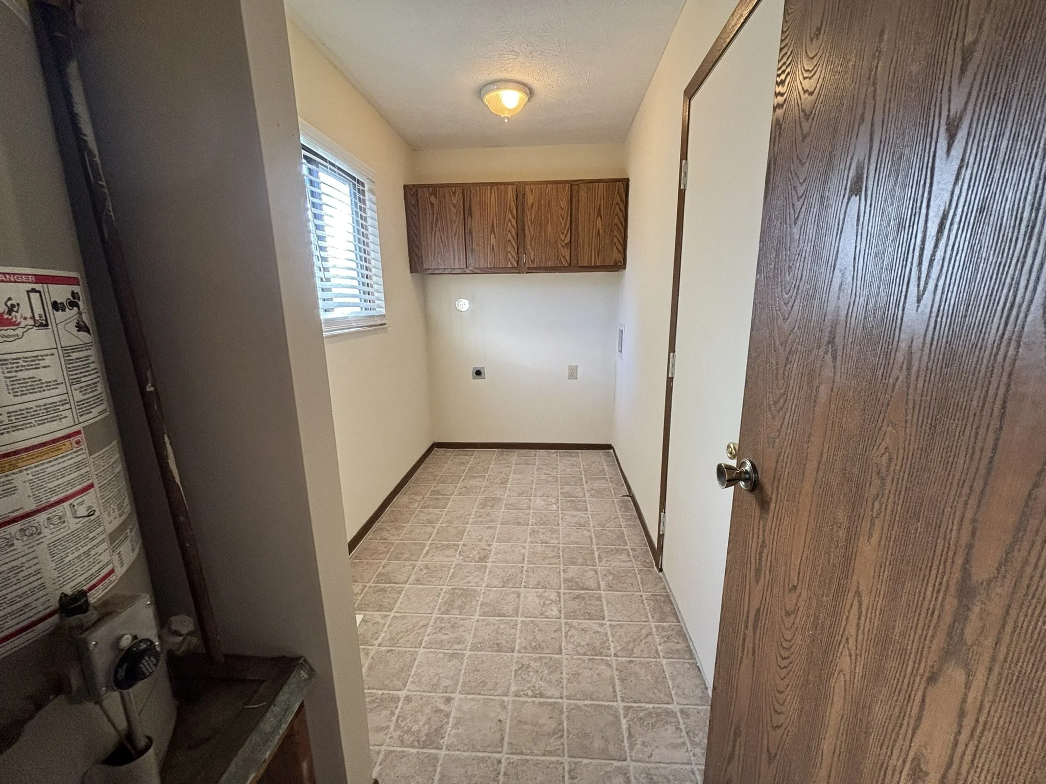 Empty laundry or utility room with beige tile flooring, a window with blinds, a wooden cabinet on the far wall, and a door on the right. Part of a water heater is visible on the left.