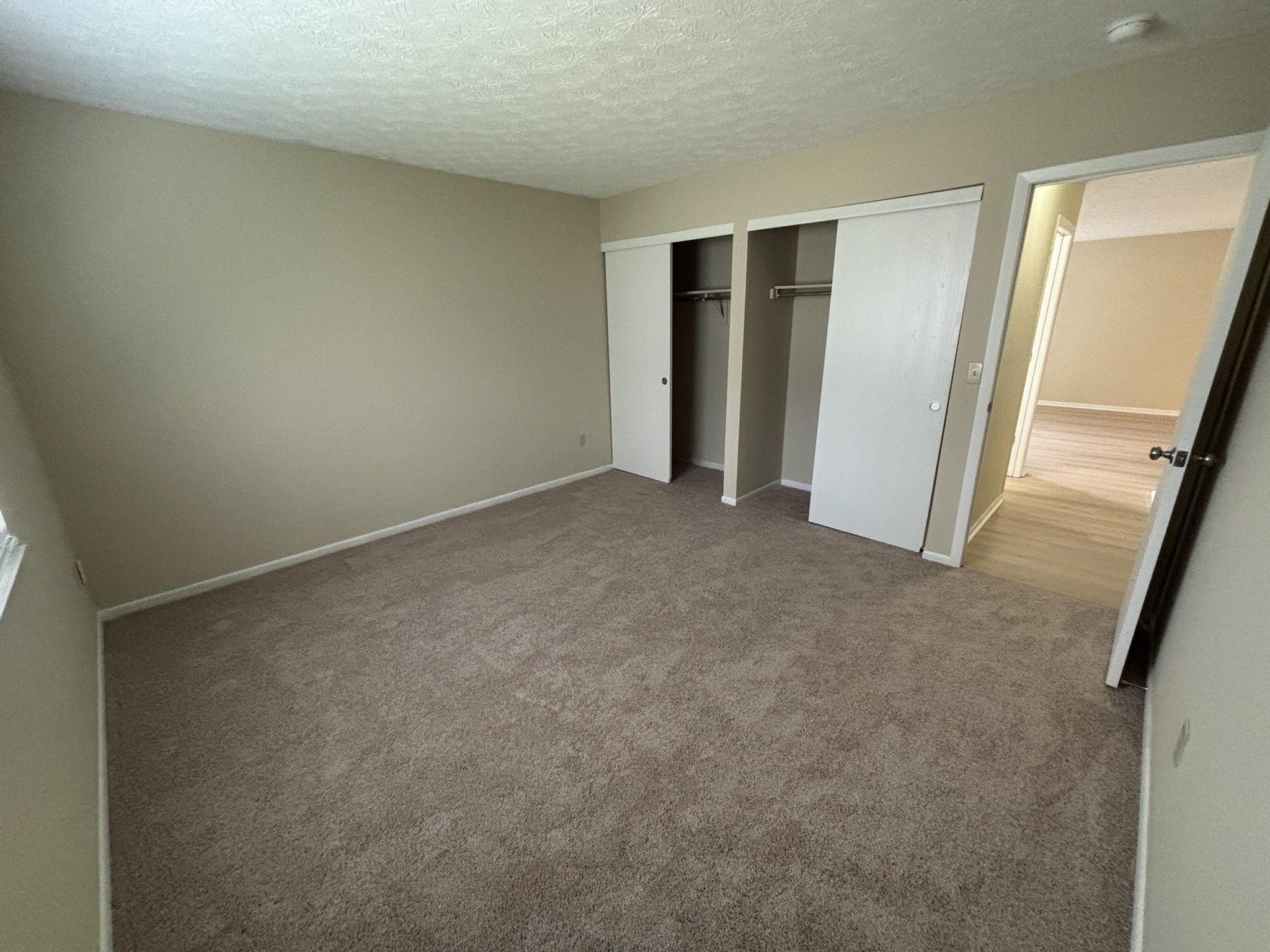 Empty bedroom with beige walls, brown carpet, an open closet with sliding doors, and a doorway leading to an adjacent room with light-colored flooring.