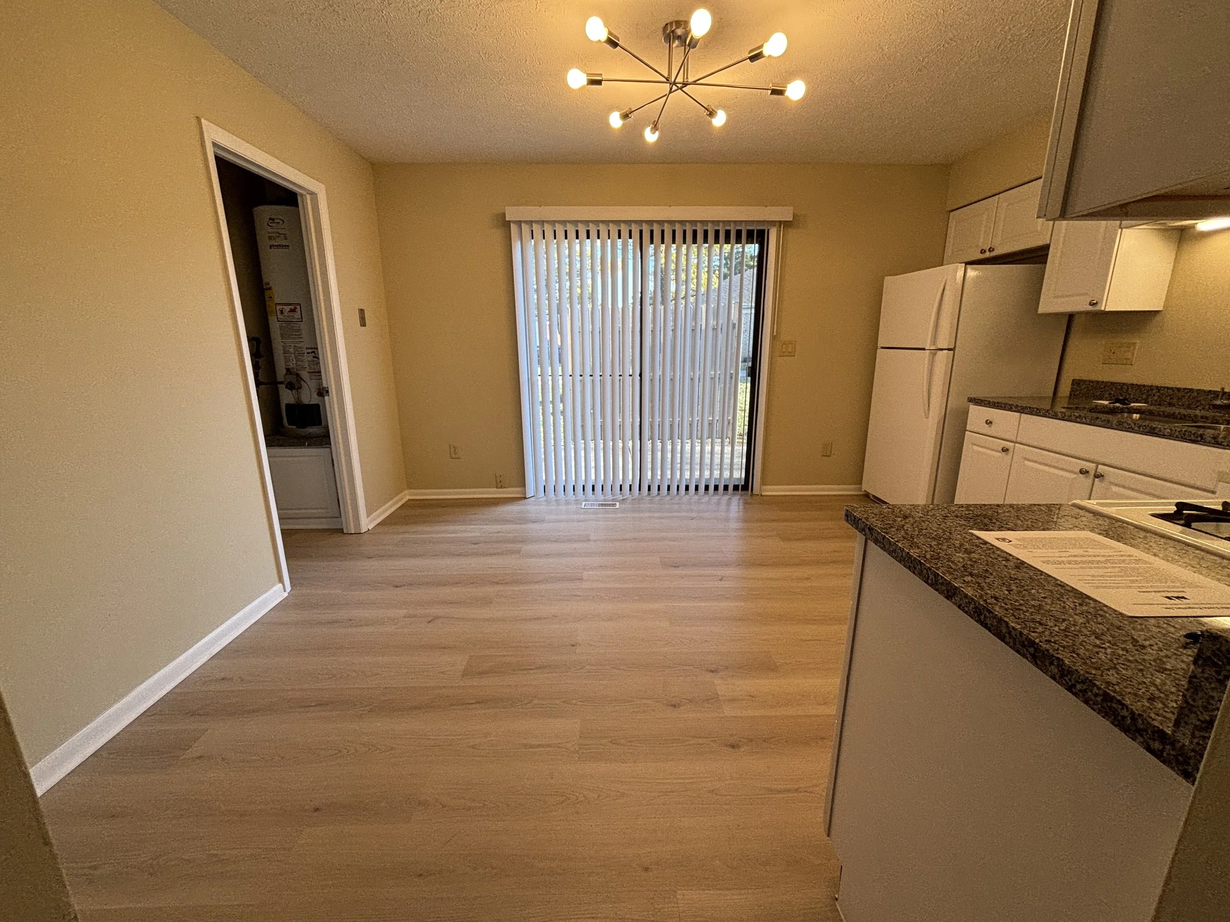 Empty kitchen and dining area with hardwood flooring, white cabinets, granite countertops, a refrigerator, a sliding glass door with vertical blinds, and a modern chandelier.