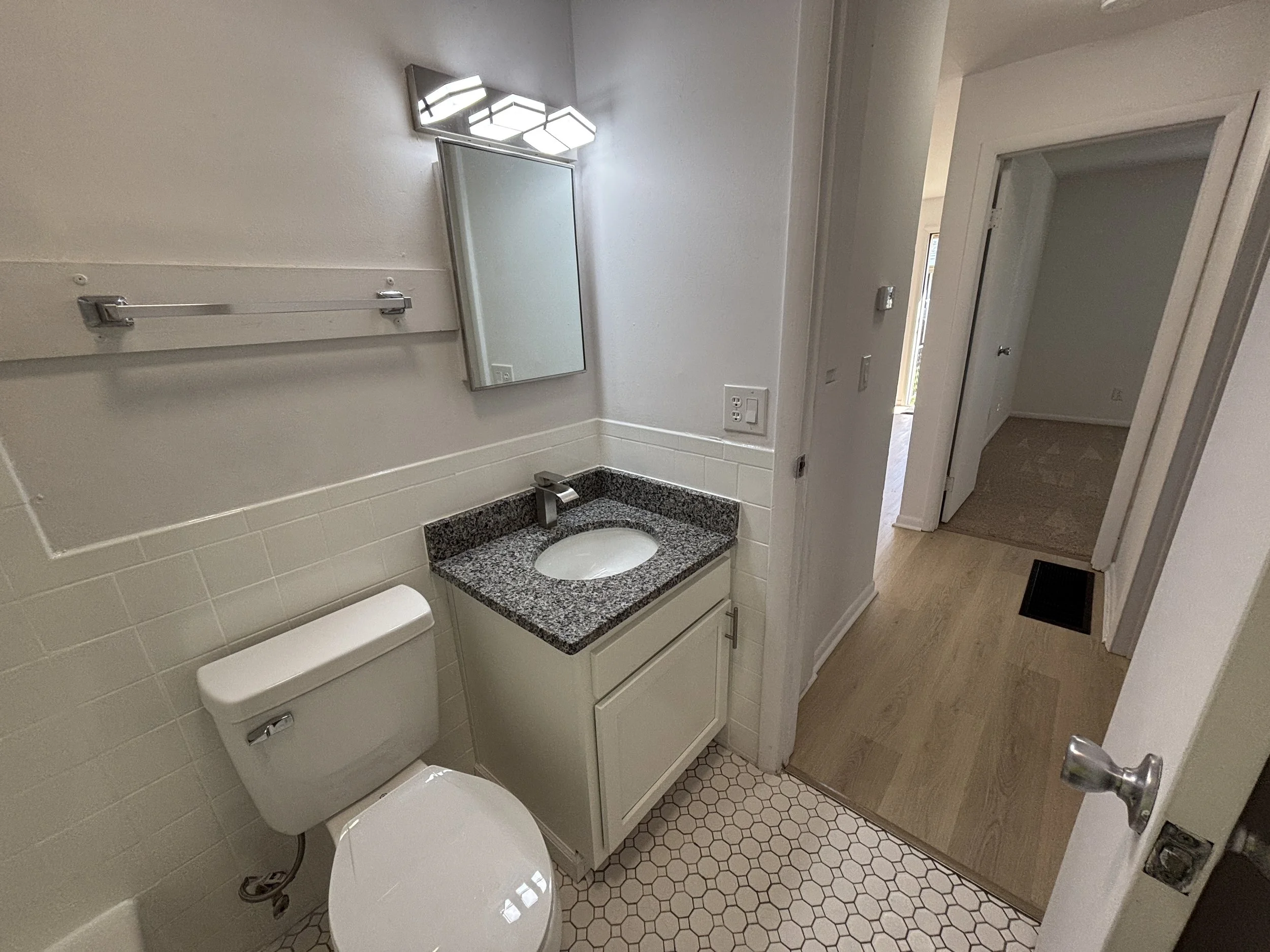 A small bathroom with a white toilet, a beige vanity with a granite countertop and oval sink, a mirror, and a three-panel light fixture. The floor has small hexagonal tiles, and the hallway outside shows a wooden floor.