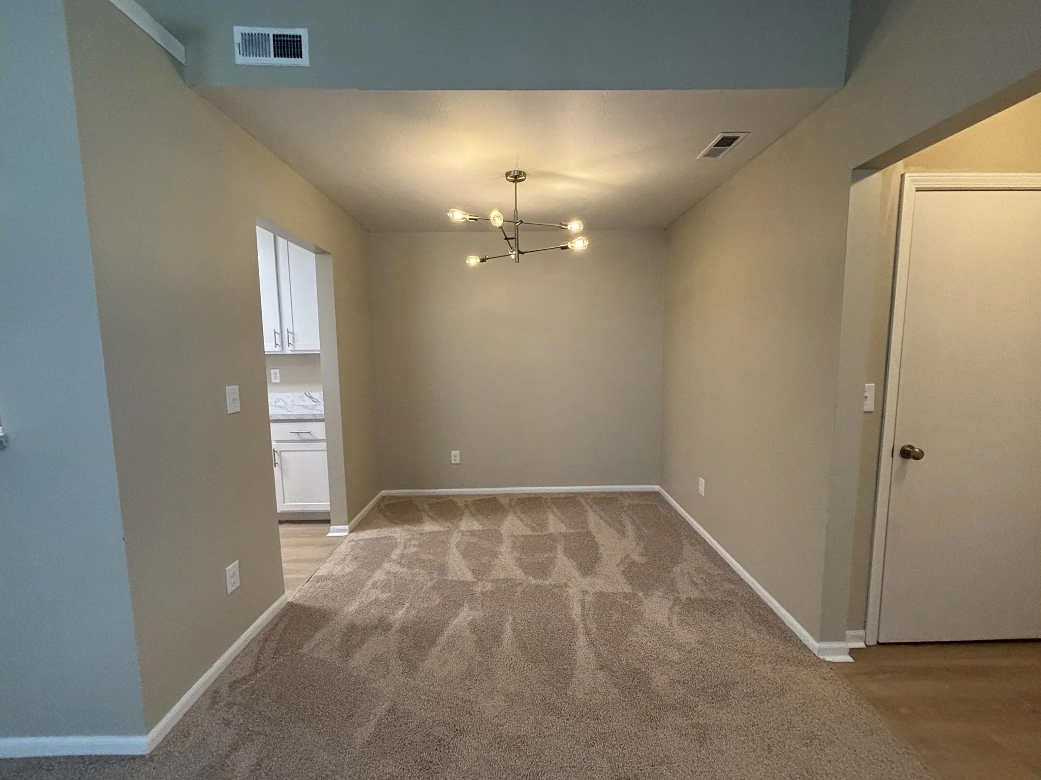 Empty dining area with a modern chandelier, beige walls, carpeted floor, and an adjacent kitchen visible through a doorway.