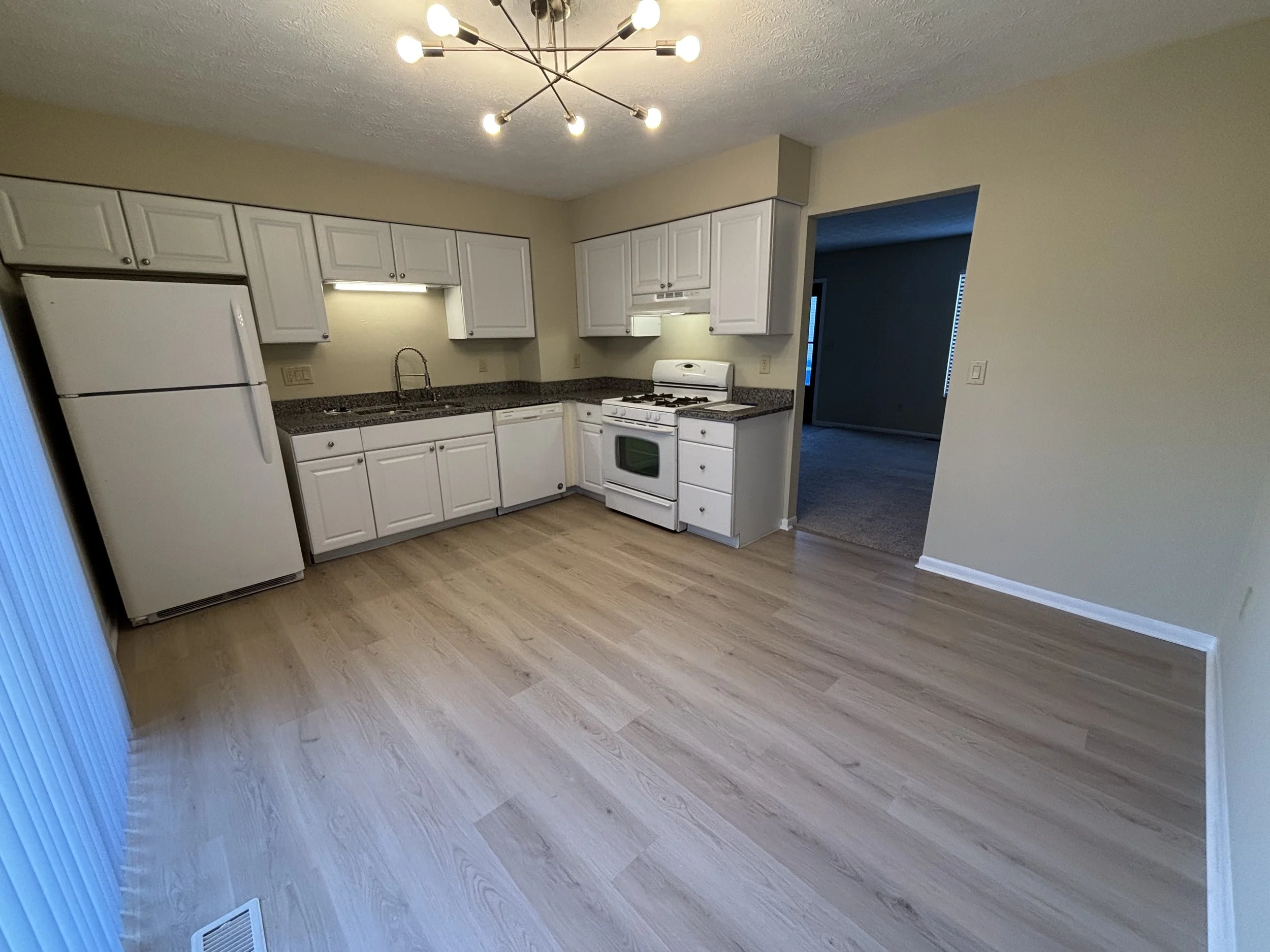 Empty kitchen with white cabinets, a white refrigerator, a white stove, granite countertops, a stainless steel sink, and light-colored wood flooring. A modern chandelier hangs from the ceiling.