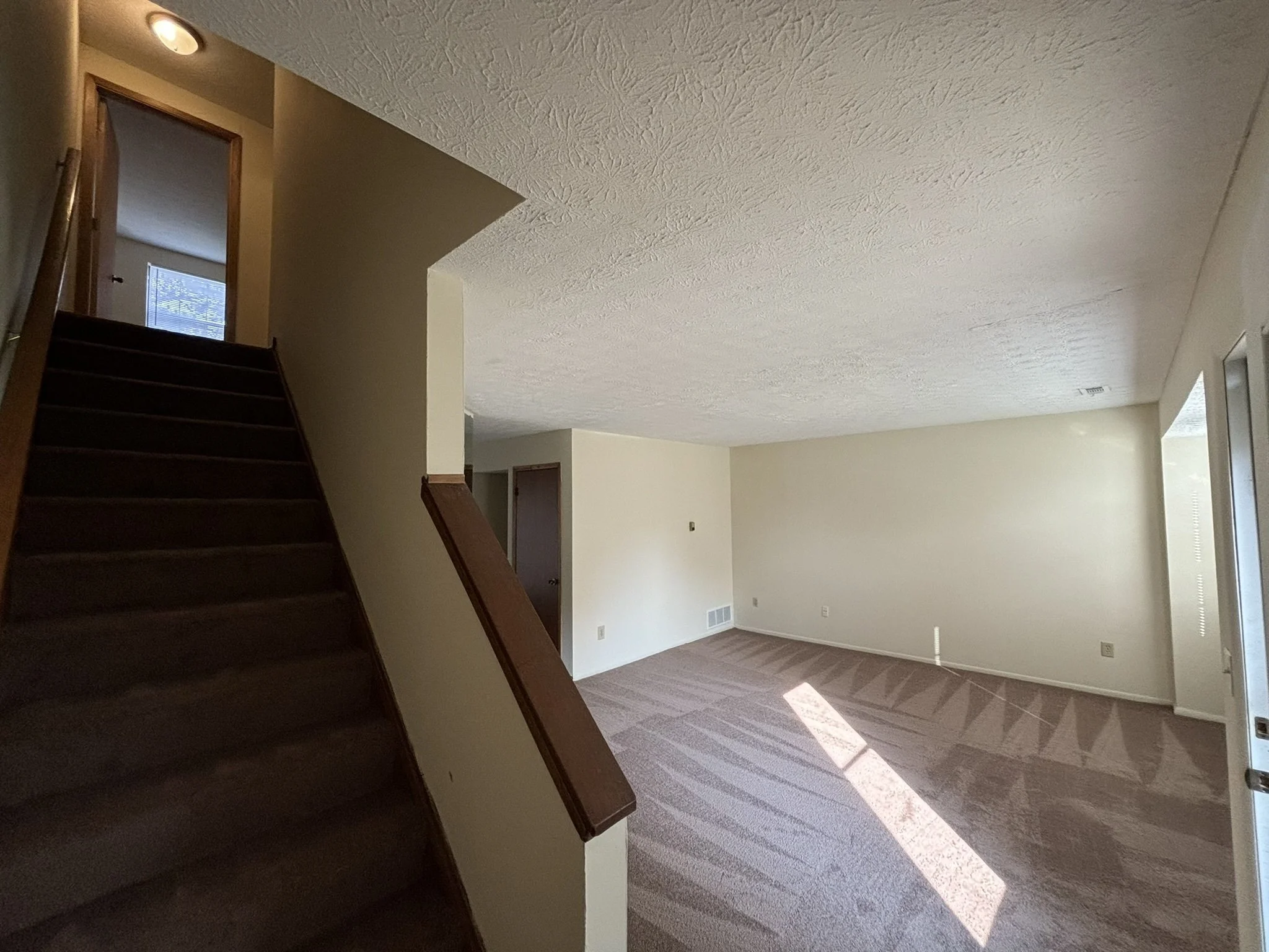 Empty living room with beige carpet, white walls, a staircase with dark wood steps and railing, and a large window letting in sunlight.