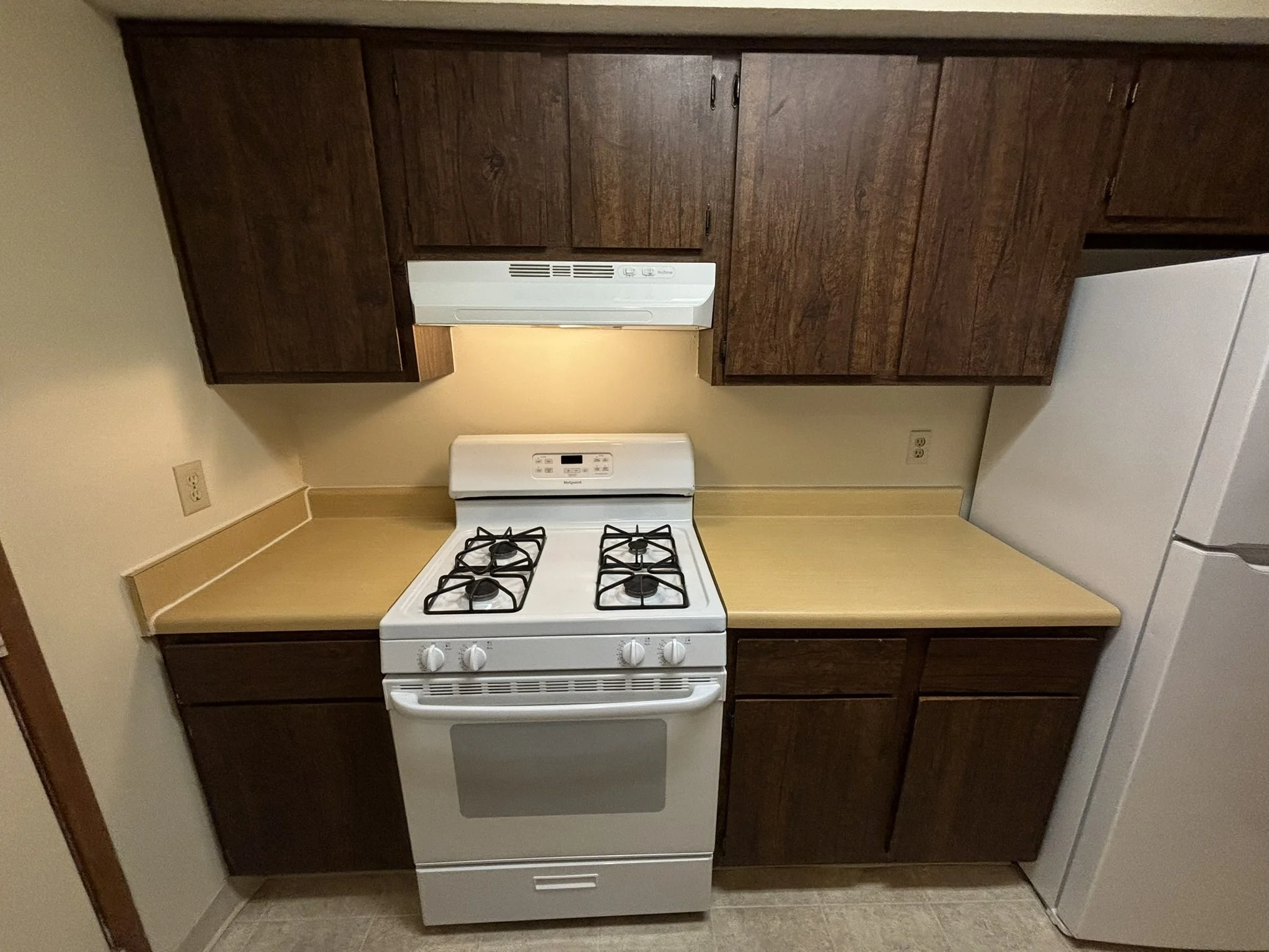 A small kitchen with dark wooden cabinets, a white gas stove, and a white refrigerator. The countertop is light-colored, and the wall behind the stove has outlets. The floor is tile.