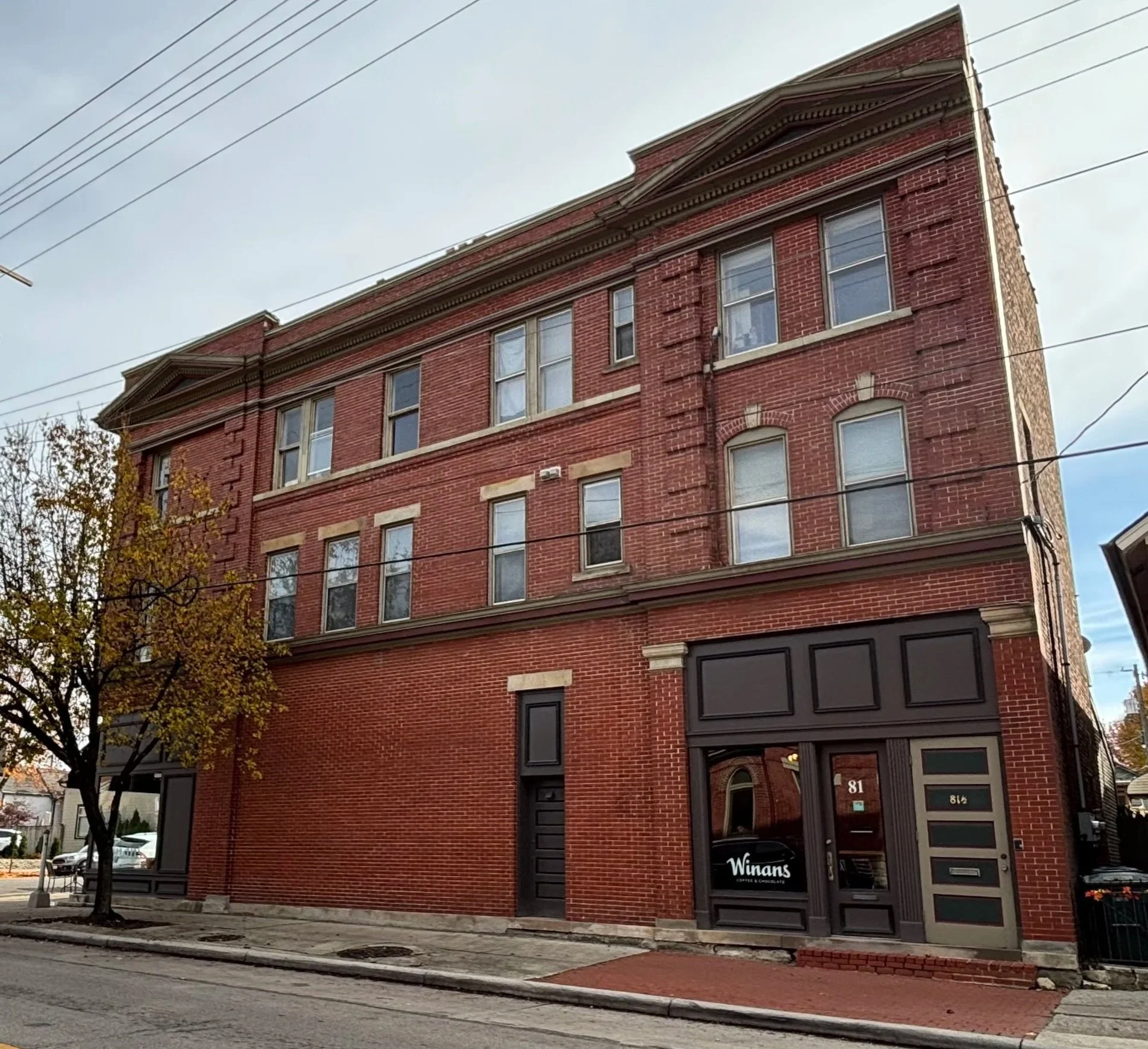 A four-story red brick building with a business called Winans on the ground floor, with signs reading 81 and 814. The building has multiple rectangular windows and decorative brickwork details, with a street and sidewalk in front, and a tree on the left side.