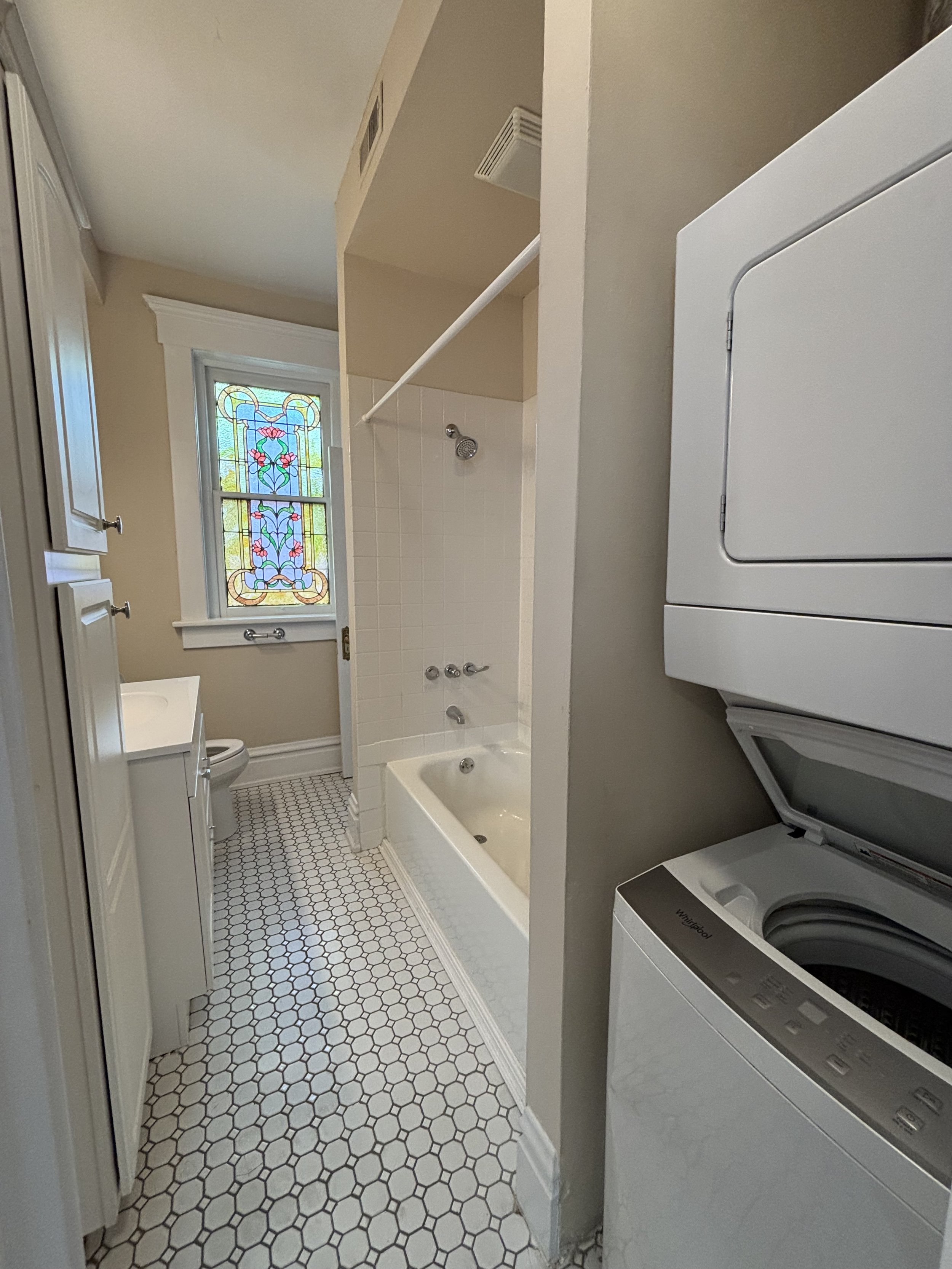 Small bathroom with a stained glass window, bathtub, toilet, washing machine, and laundry area.