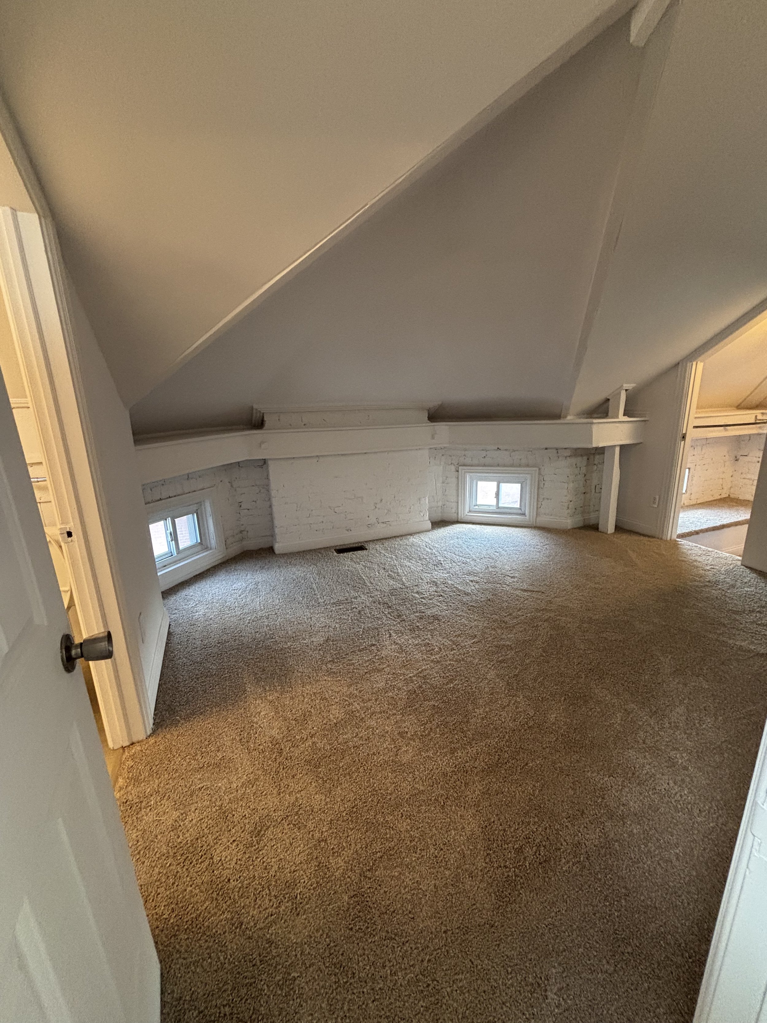 A small attic room with a sloped ceiling, white brick wall, two small windows, beige carpet, and white painted wood beams.