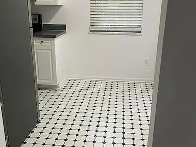 Empty kitchen with black and white checkered tile floor, white cabinets, and a window with blinds