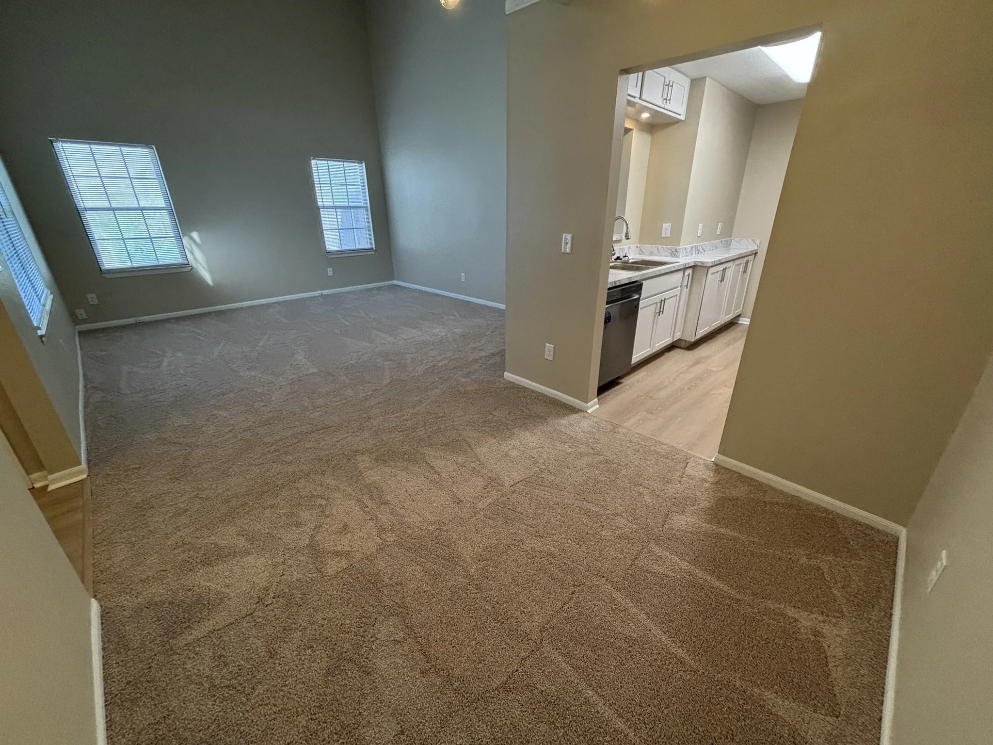 Empty living room with beige carpet, beige walls, three windows with blinds, adjacent kitchen with white cabinets and marble countertop.