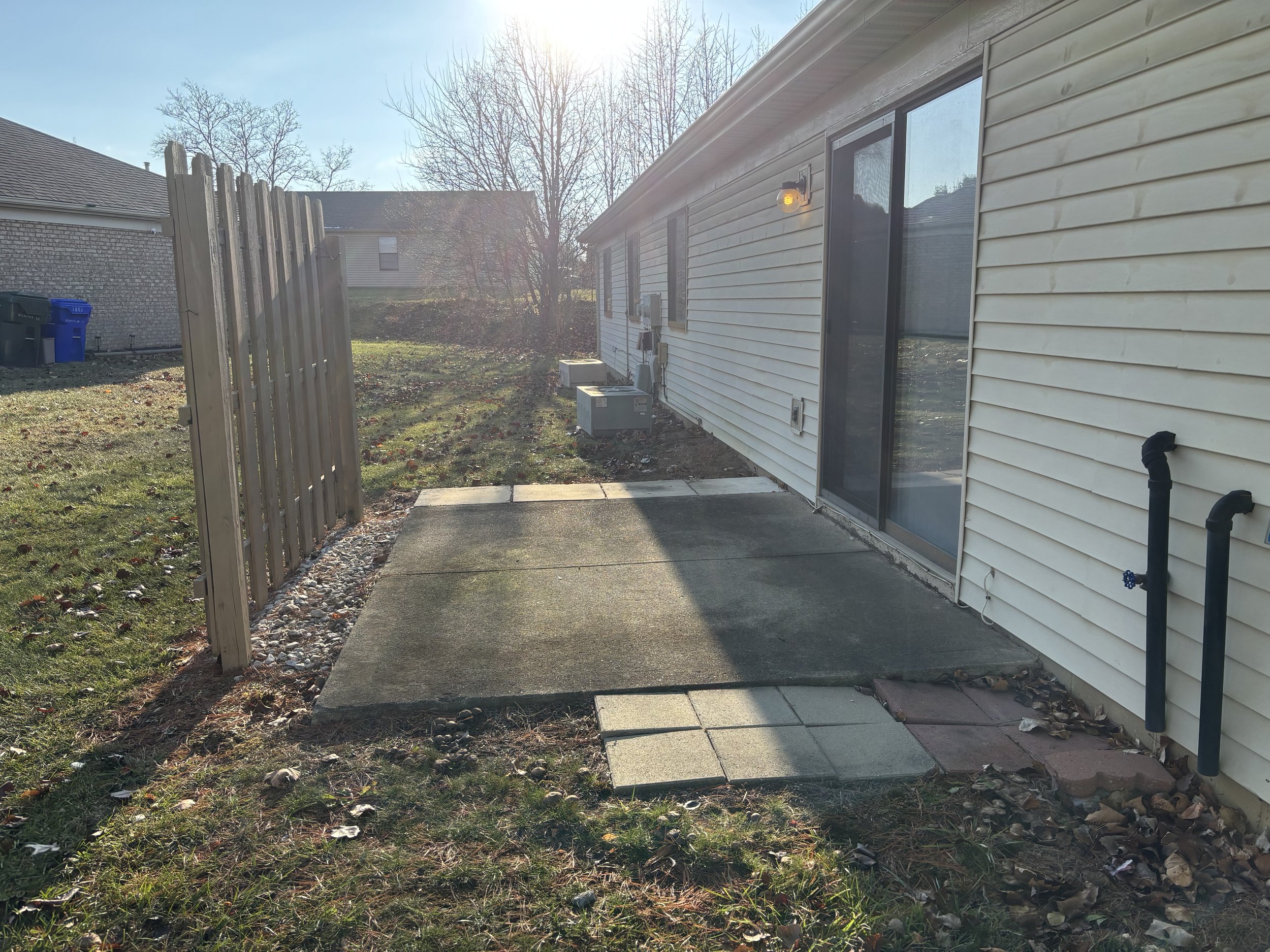 Backyard patio with concrete slabs and pavers, beige siding house with sliding glass door, black pipes on wall, wooden fence with gravel border, leaf-covered ground, and sunlight in the background.