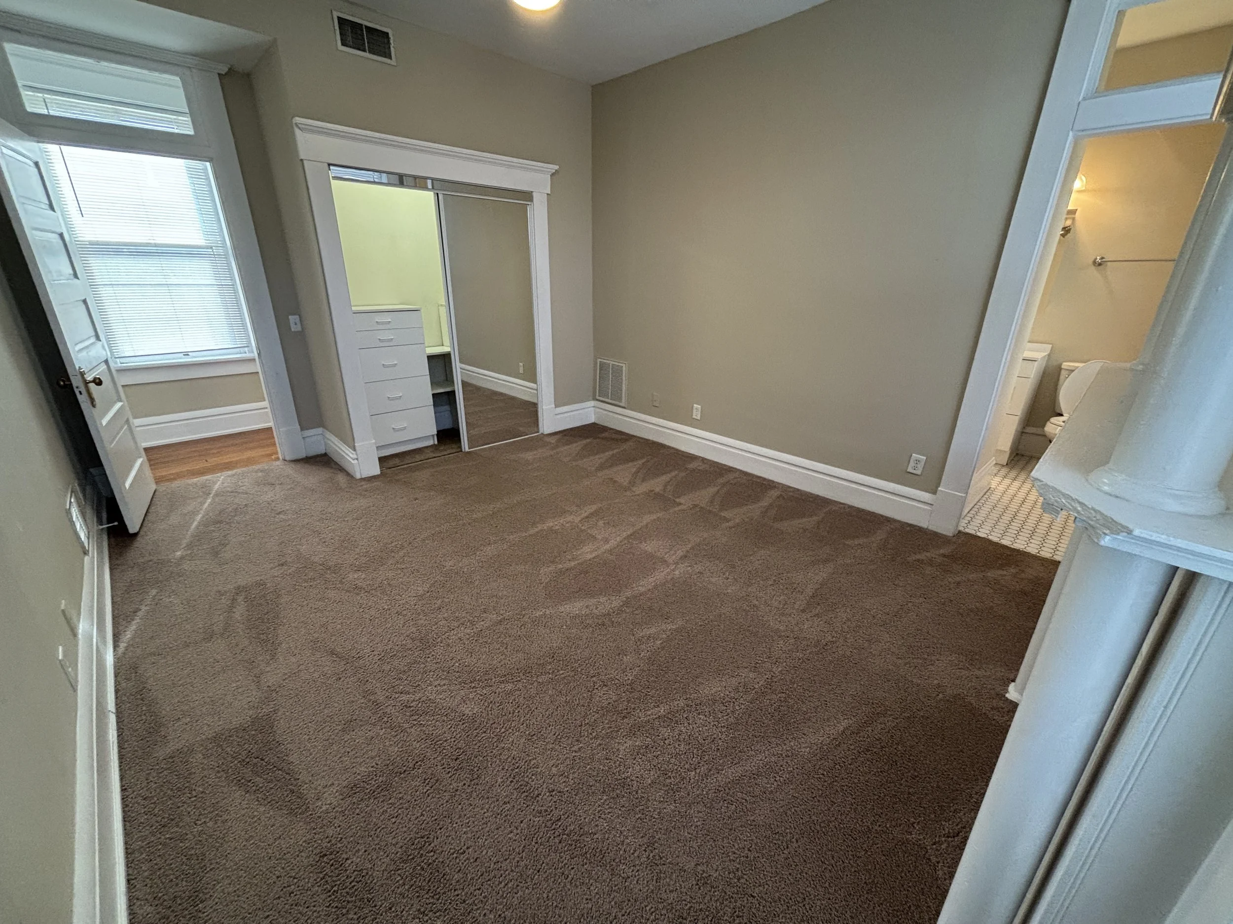 Empty living room with beige walls, brown carpet, white trim, a closet with sliding mirror doors, and a small bathroom visible through an open door on the right.