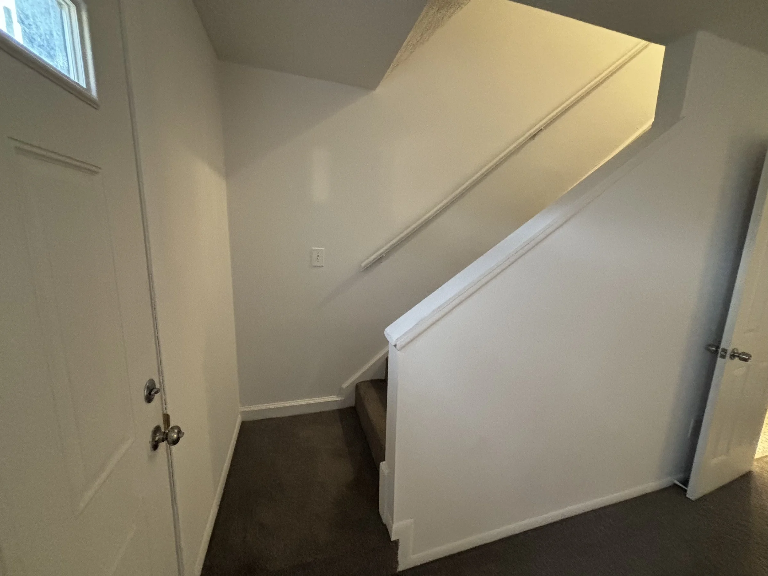Interior view of a staircase with white walls, a dark carpeted stair, a handrail, a door on the right, and a window on the left wall.