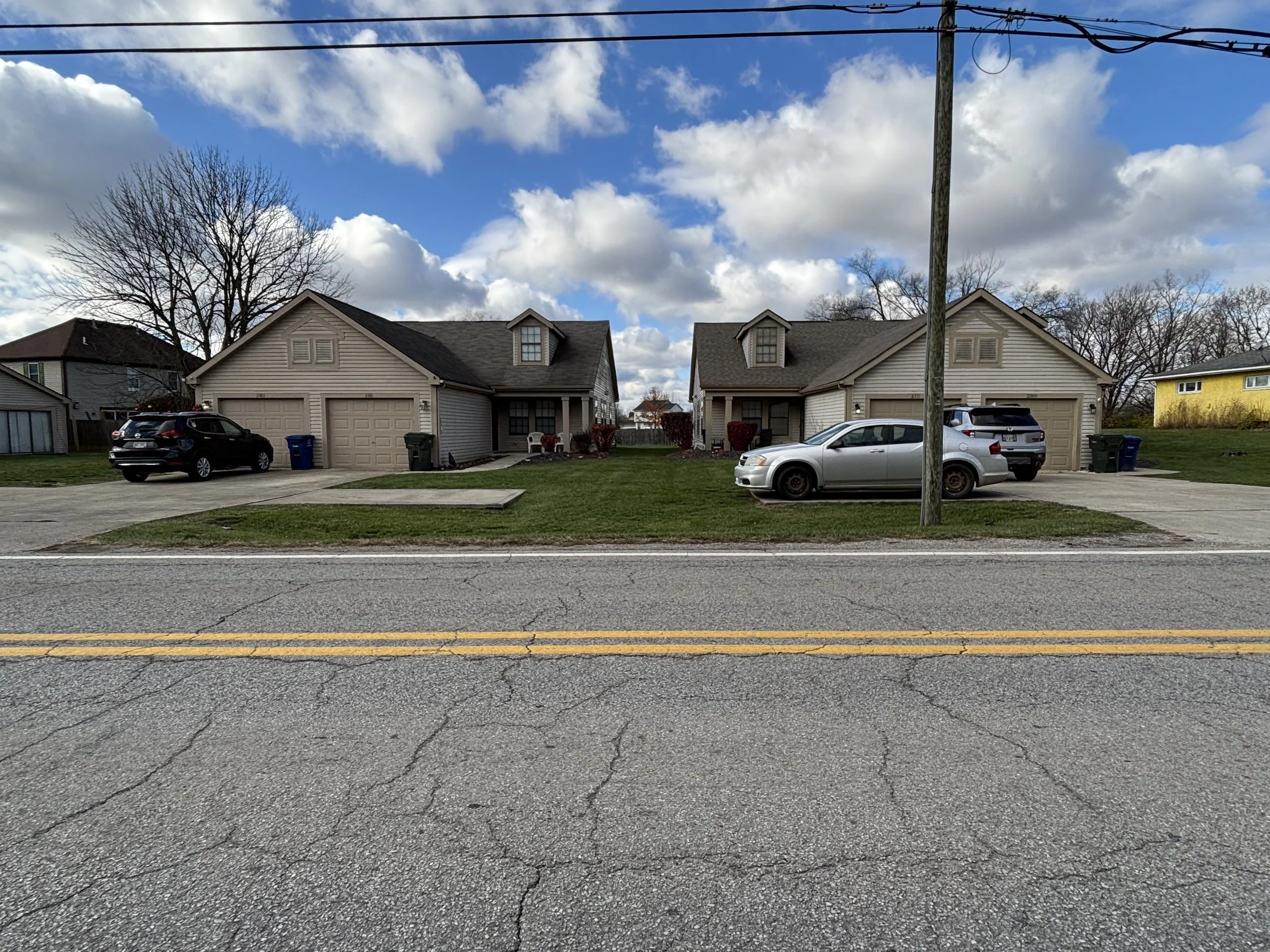 Two beige residential duplexes with attached garages and front lawns, parked cars, a cable pole, and a paved street with yellow lines under a partly cloudy sky.
