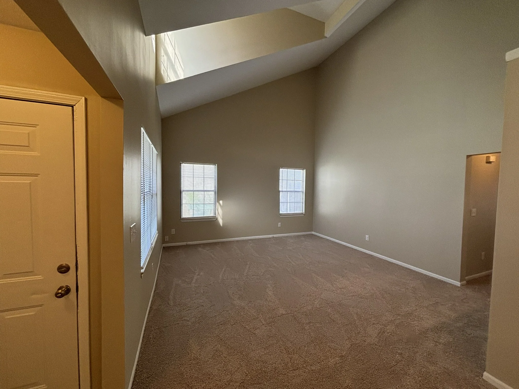 Empty living room with beige carpet, cream-colored walls, three windows with blinds, and a high, vaulted ceiling.