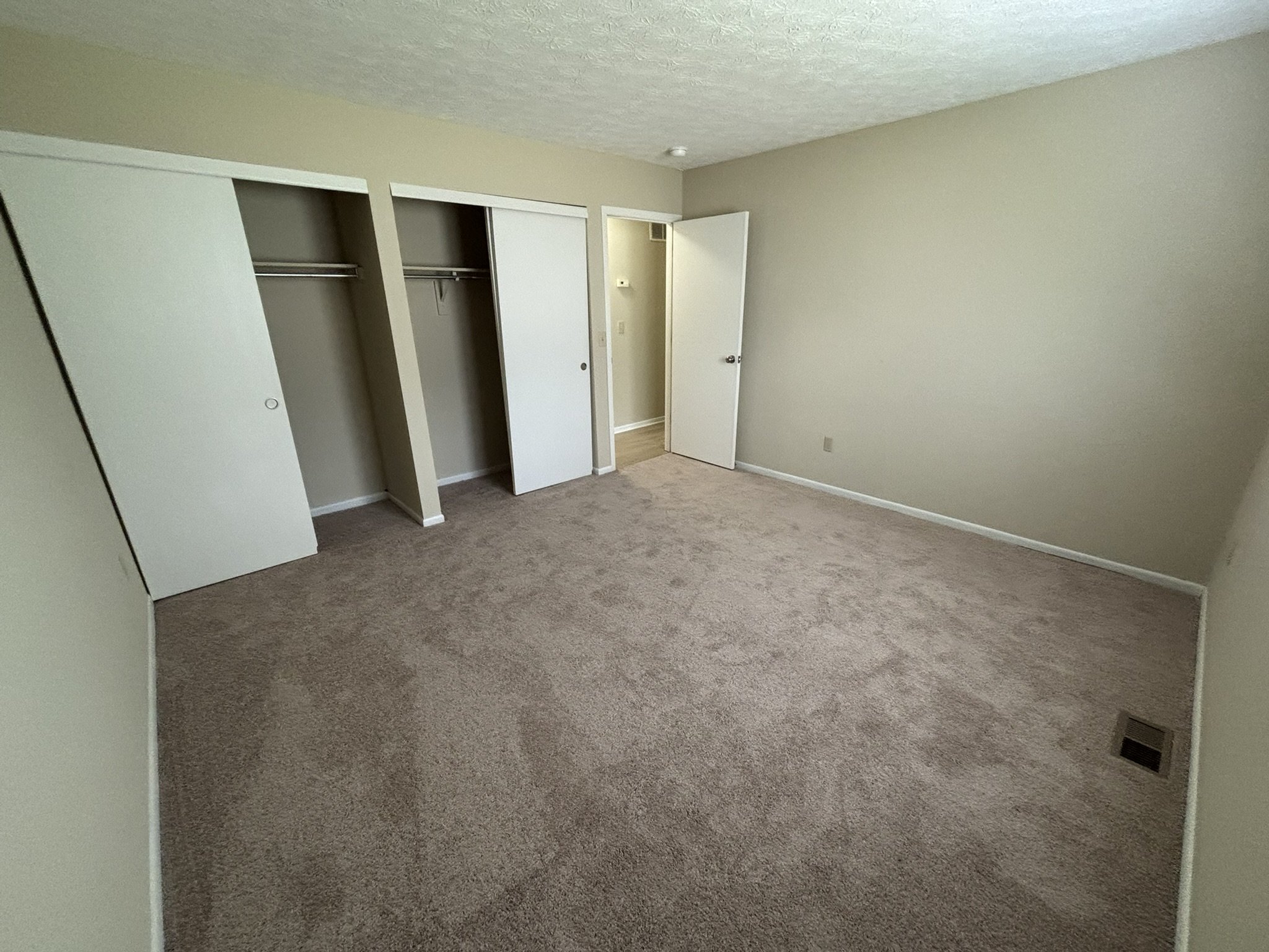 Empty bedroom with beige carpet, beige walls, an open closet with sliding doors, and a small hallway visible through an open door.