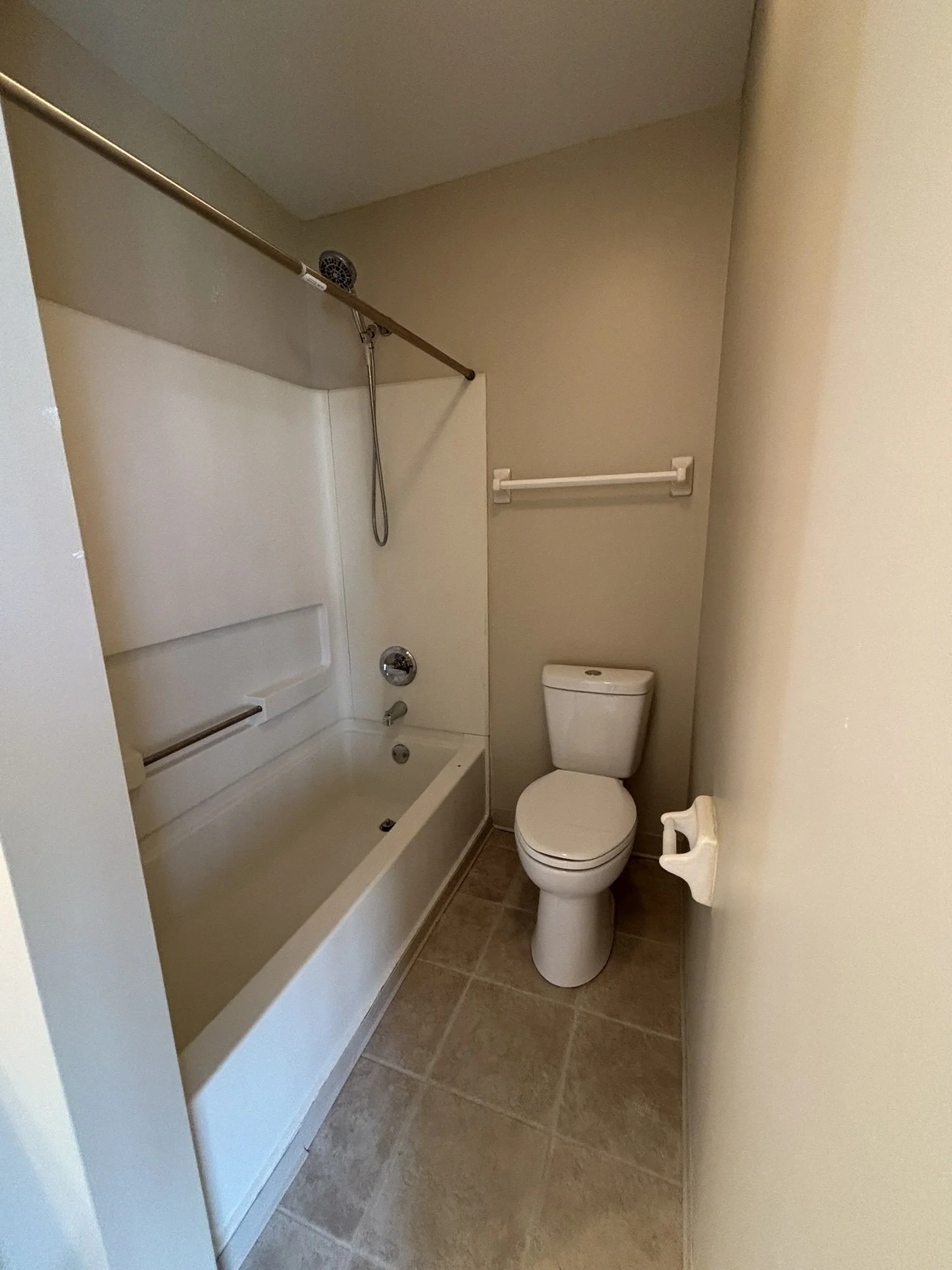 Small bathroom with a shower-bathtub combo on the left, a toilet on the right, and a beige wall with a white towel bar above the toilet.