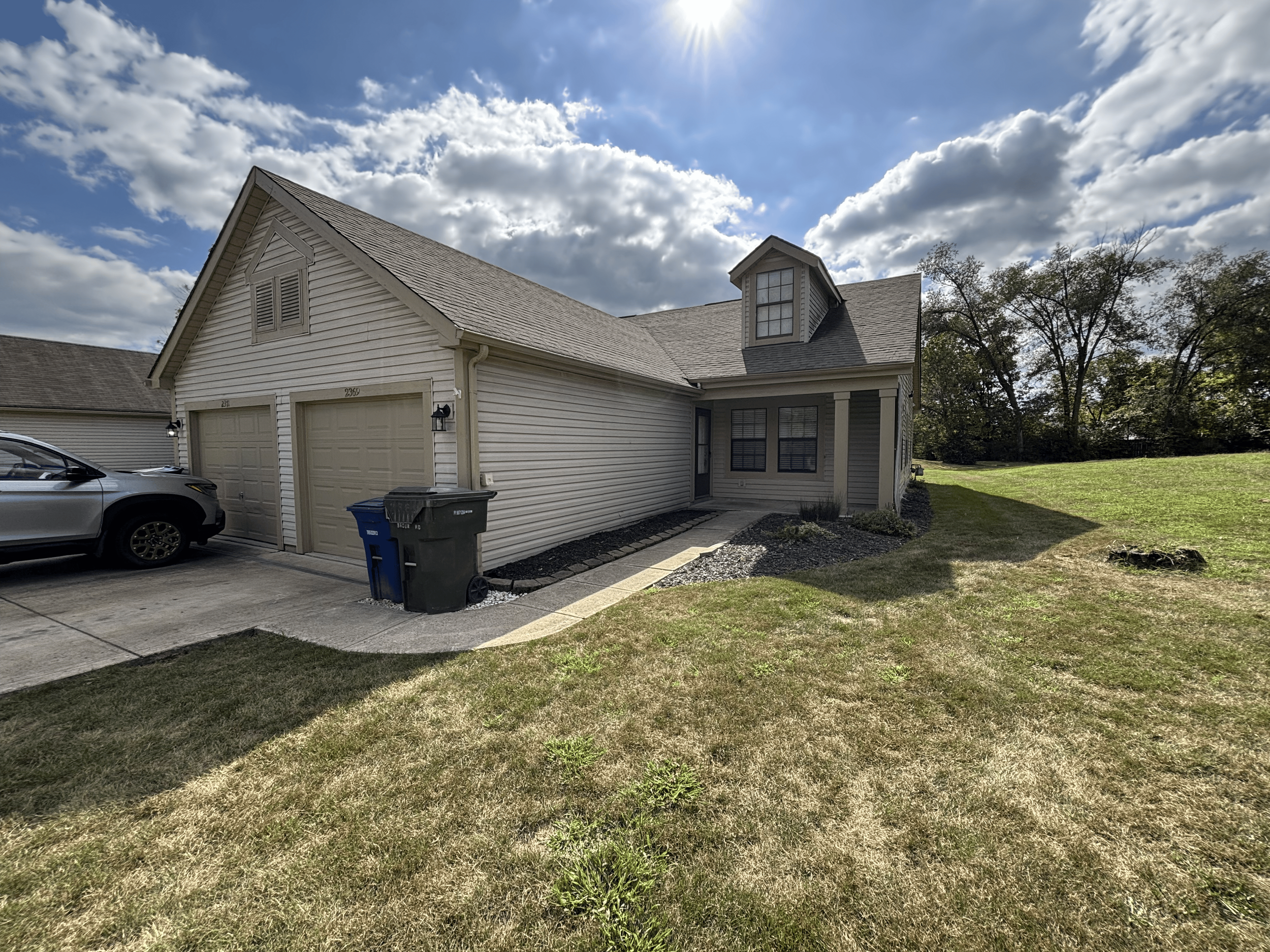 A suburban house with a beige garage door, a silver SUV parked in the driveway, and a neatly maintained lawn with a walkway leading to the front door. The house has beige siding, a small dormer window, and some trees in the background. The sky is par