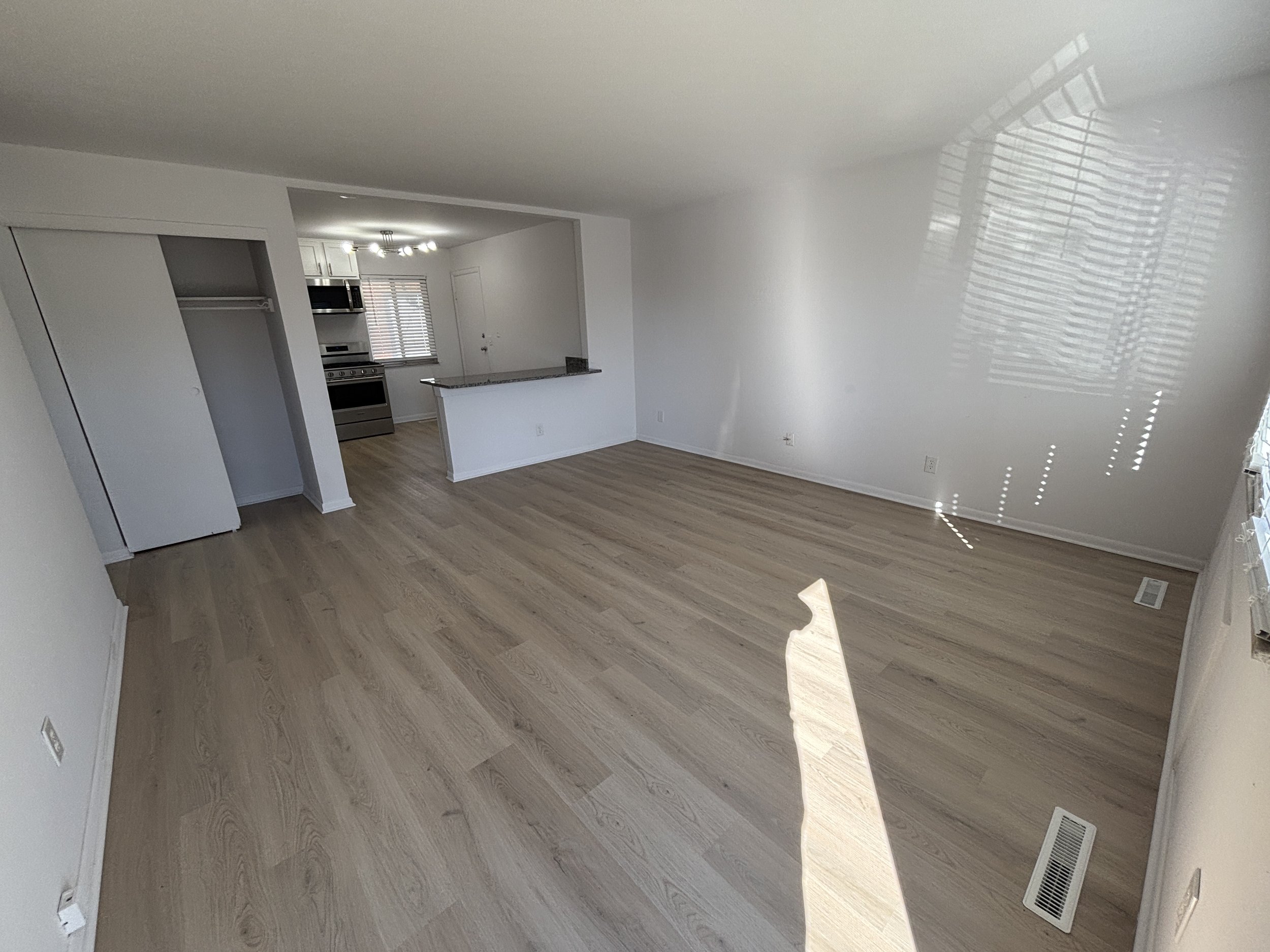 Empty living room with light wood flooring, white walls, and sunlight through window blinds, with a view of the kitchen in the background.
