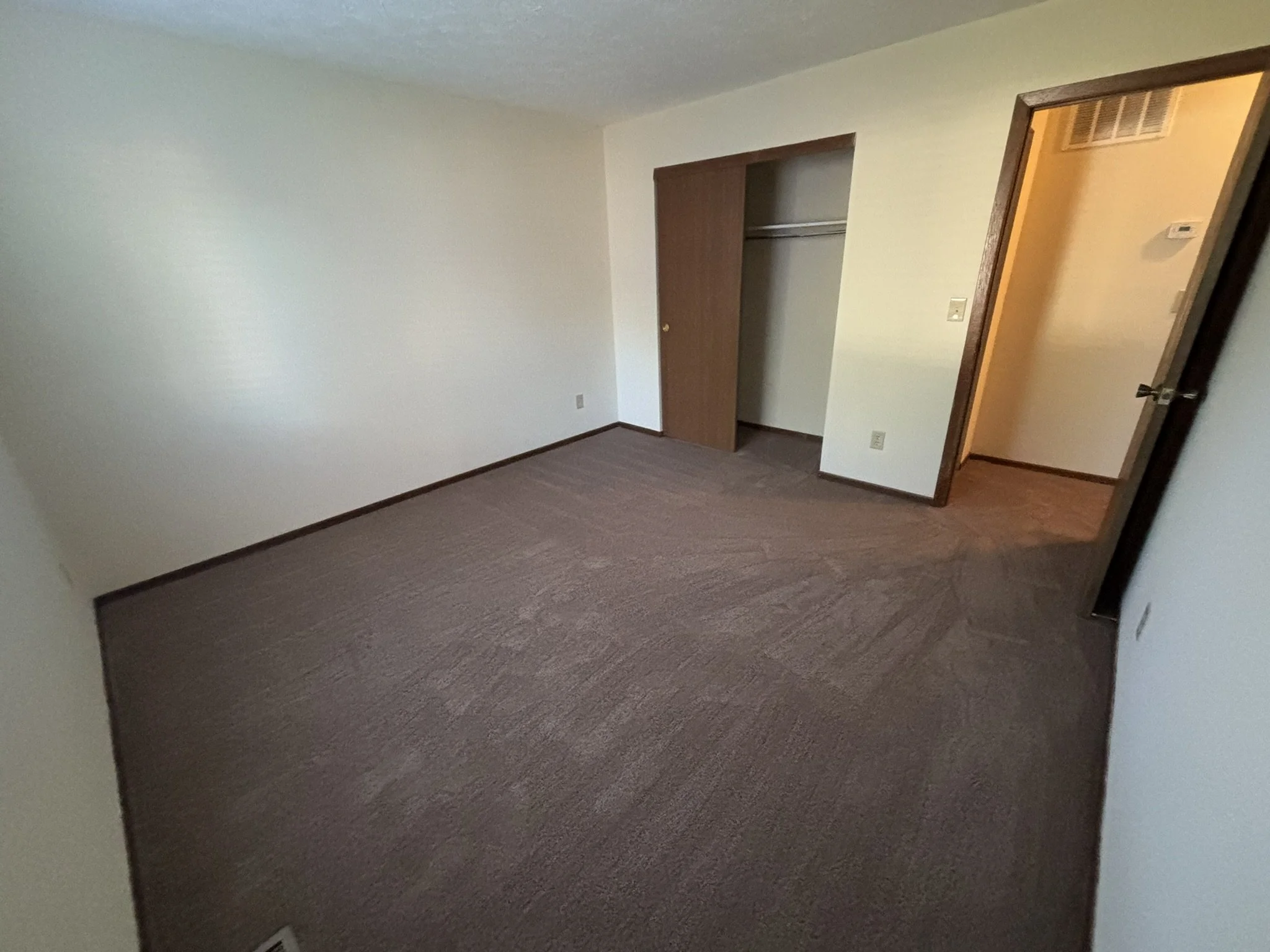 An empty bedroom with beige carpet, white walls, an open closet with a wooden sliding door, and an open door leading to a hallway.