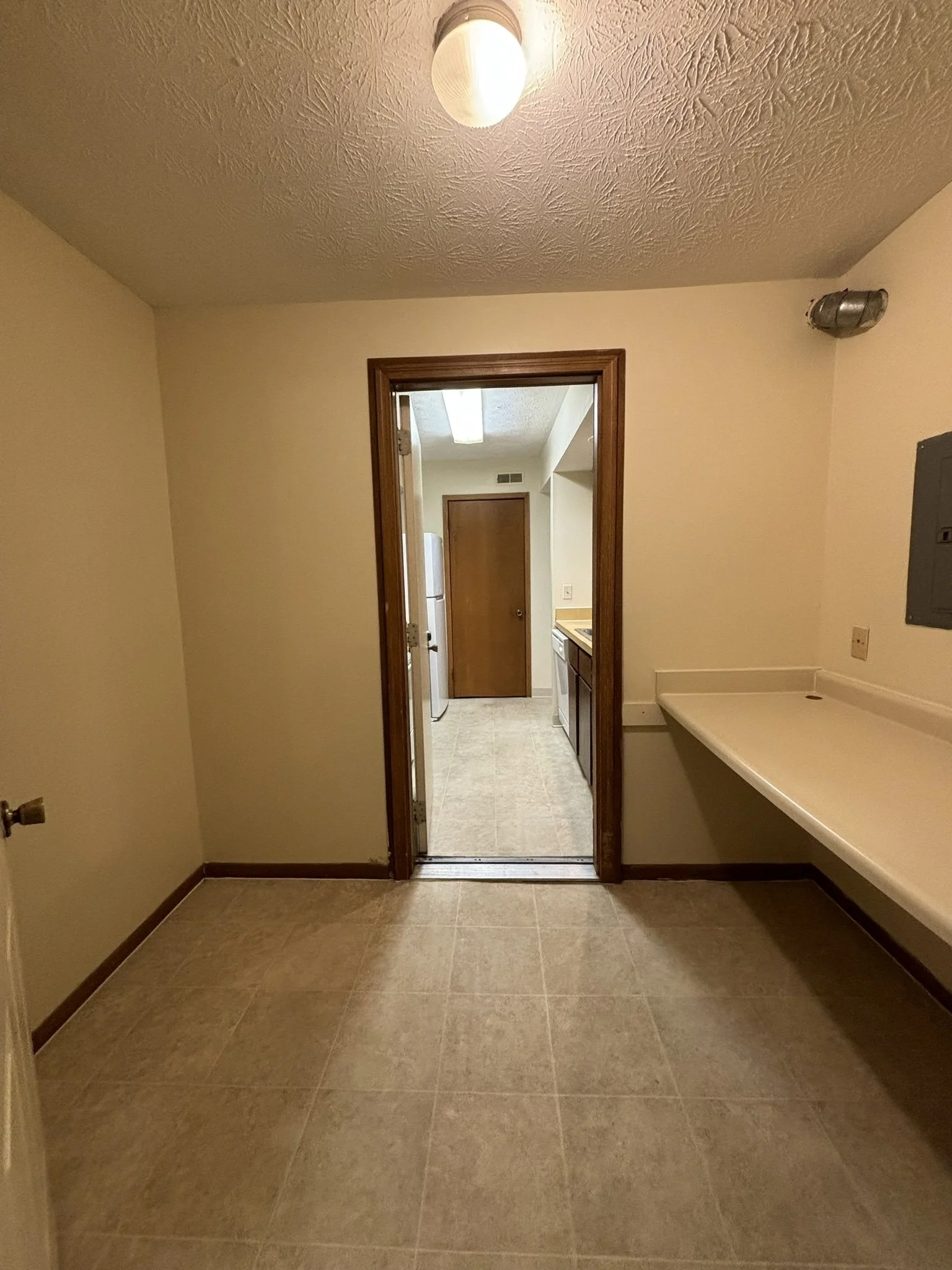 Empty room with beige tiled floor, beige walls, ceiling light fixture, electrical panel on the right wall, and a doorway leading to a kitchen with a refrigerator, counter, and wooden cabinets.