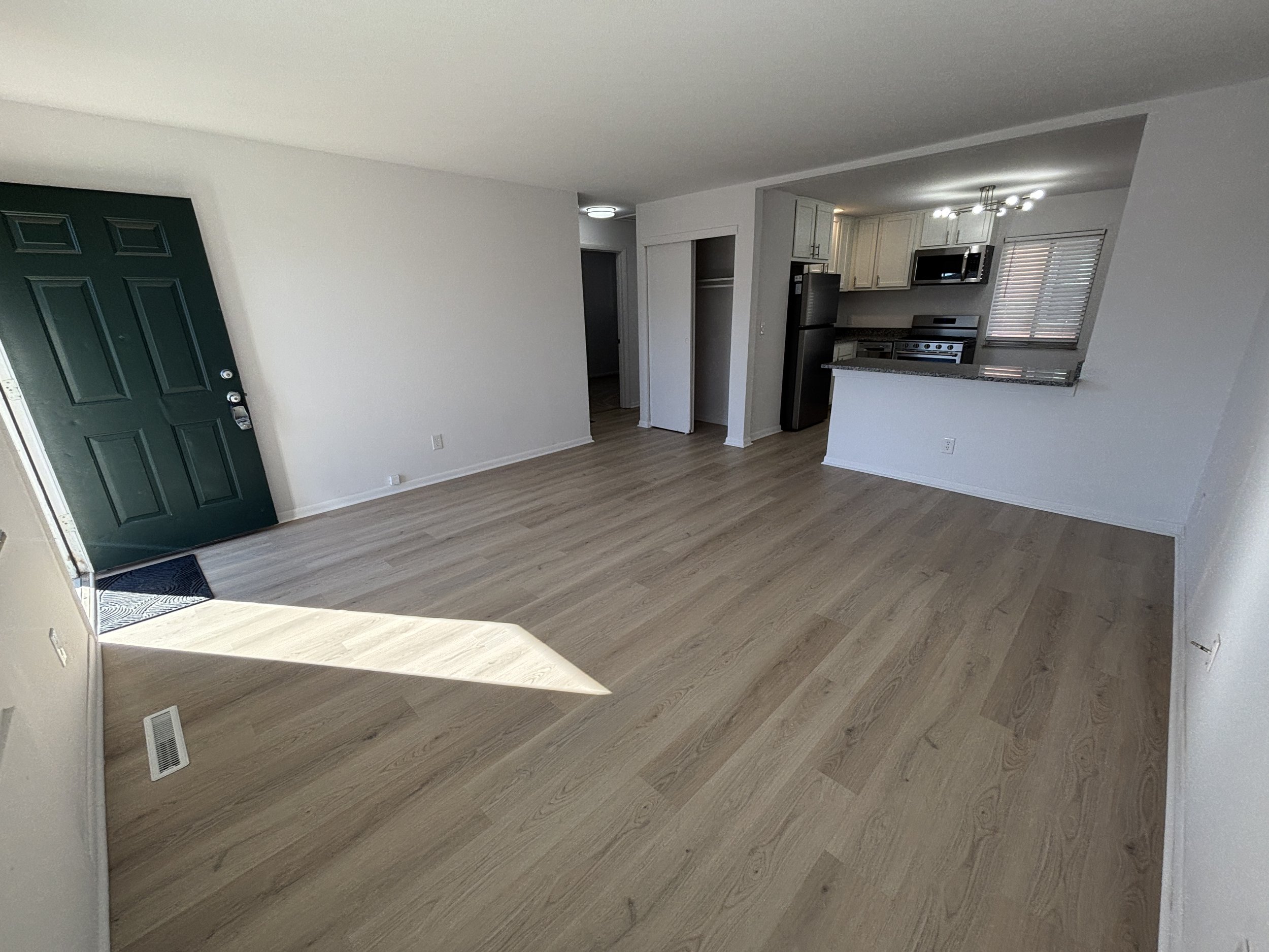 Empty living room with light wood floors, green front door, white walls, and an open kitchen with white cabinets and stainless steel appliances.
