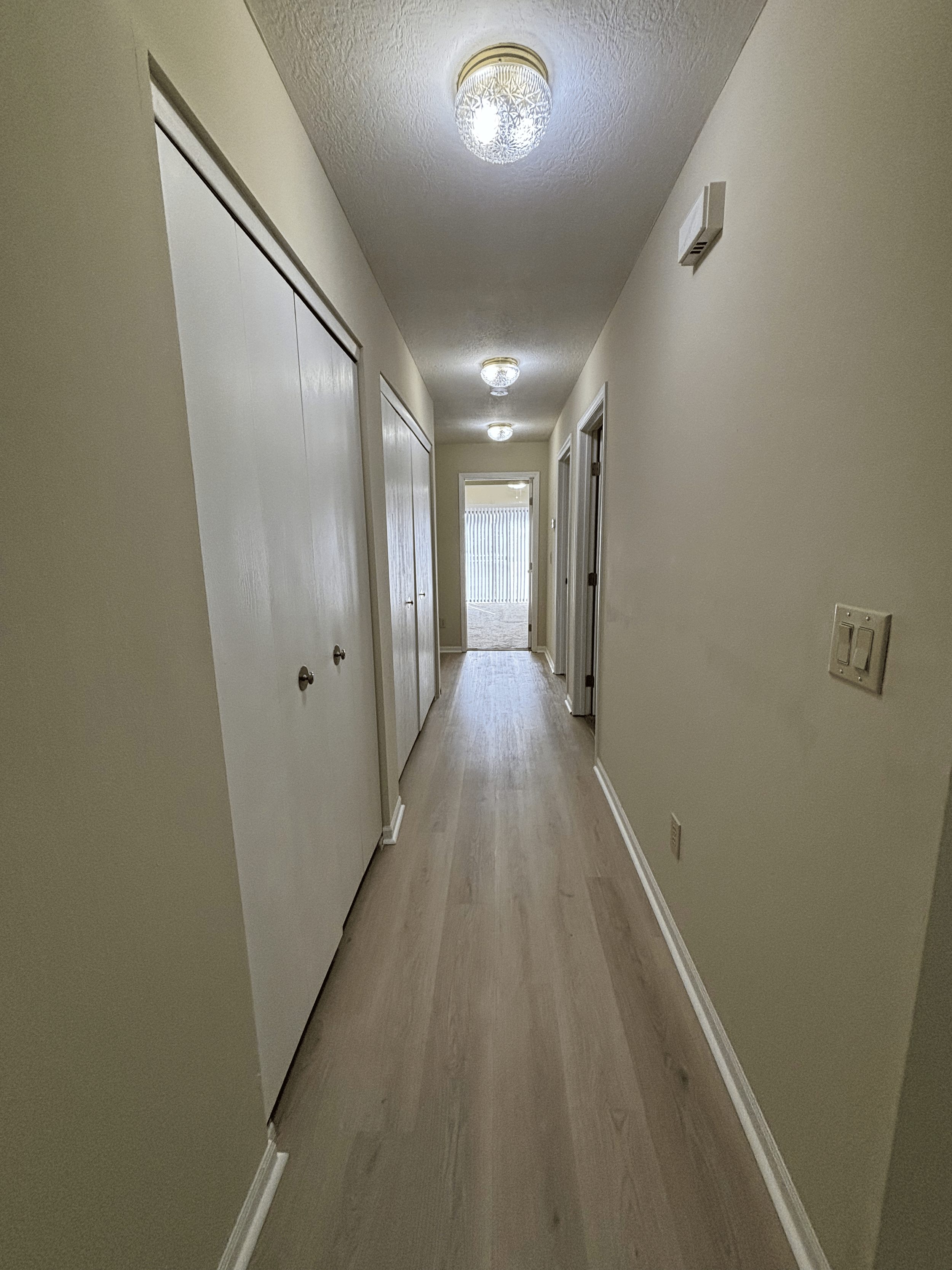 Empty hallway with wooden flooring, beige walls, ceiling lights, and closed closet doors on the left, leading to a door at the end of the hallway with a glass panel.