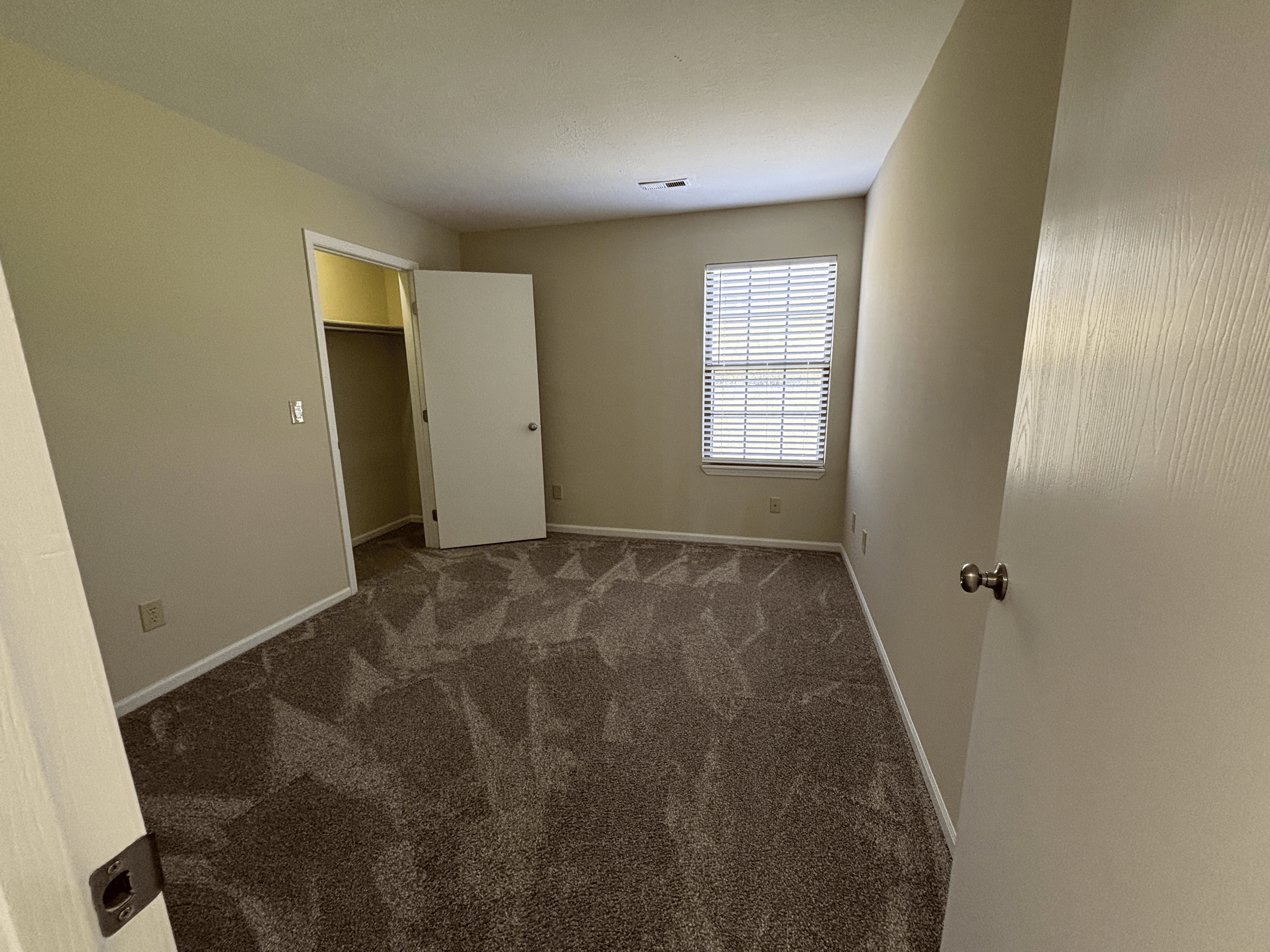 Empty bedroom with beige walls, carpeted floor, window with blinds, open closet door, and door to the room slightly ajar.