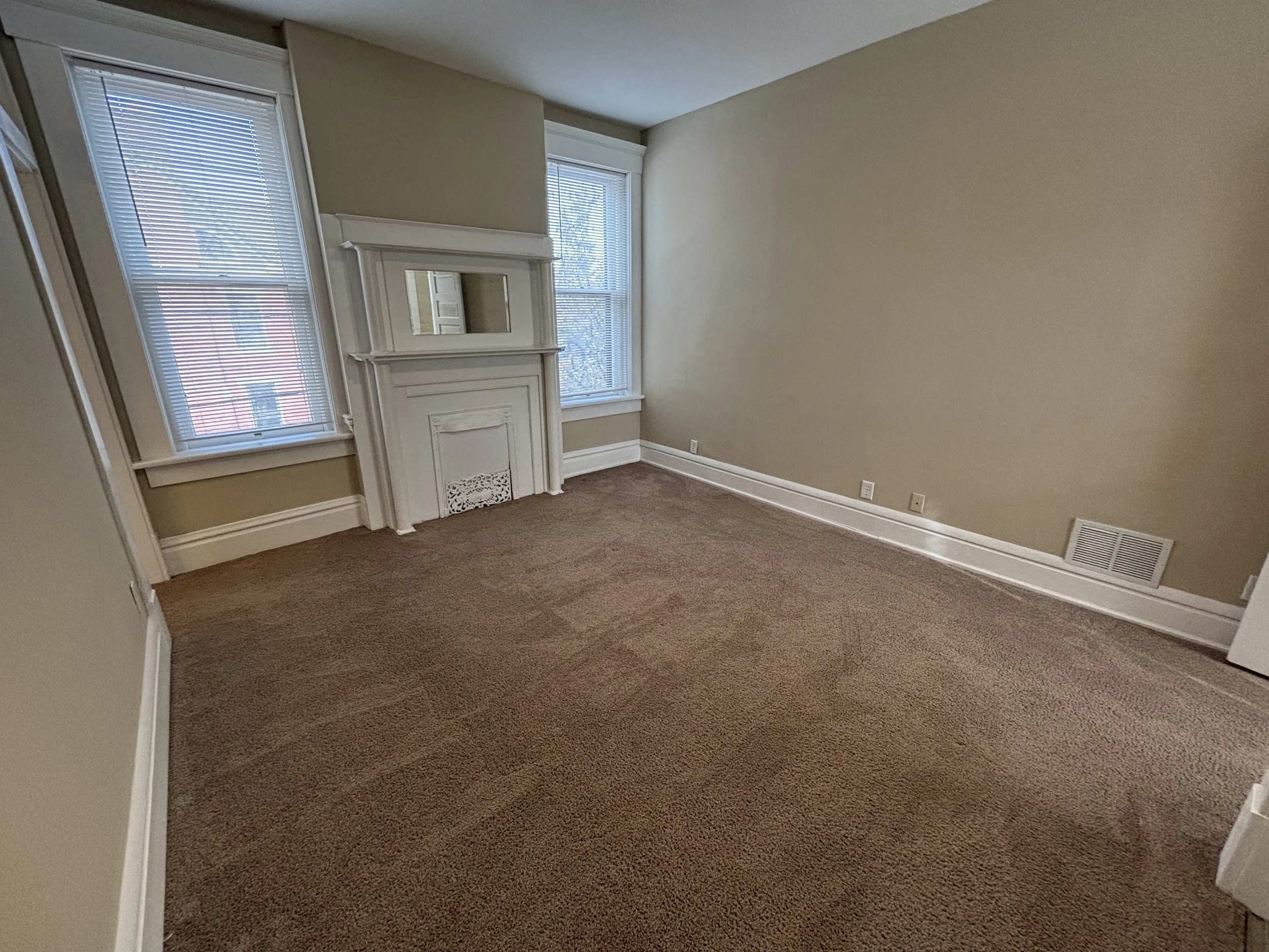 Empty room with beige walls, brown carpet, two windows with blinds, a decorative white fireplace with a mirror above, and a vent on the wall near the floor.