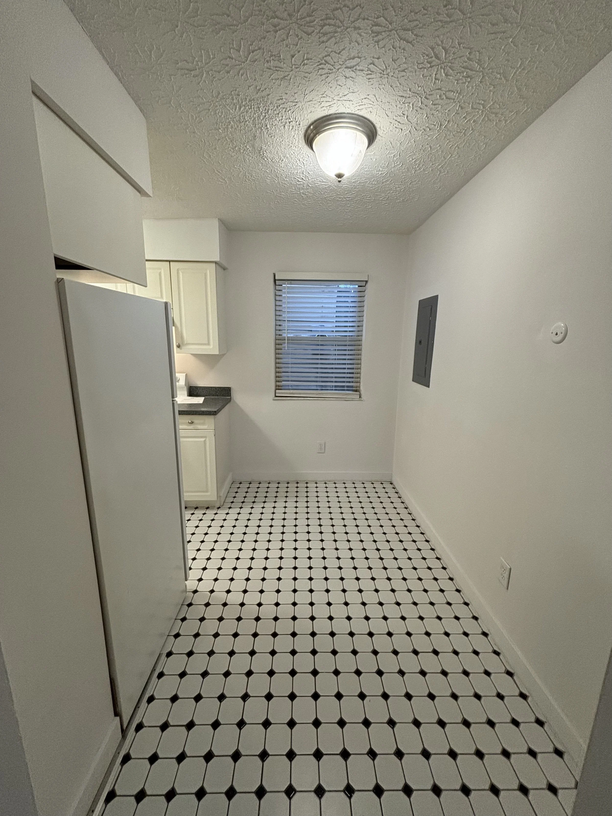 Empty kitchen with white cabinets, a window with blinds, black and white patterned tile floor, ceiling light fixture, and plain white walls.