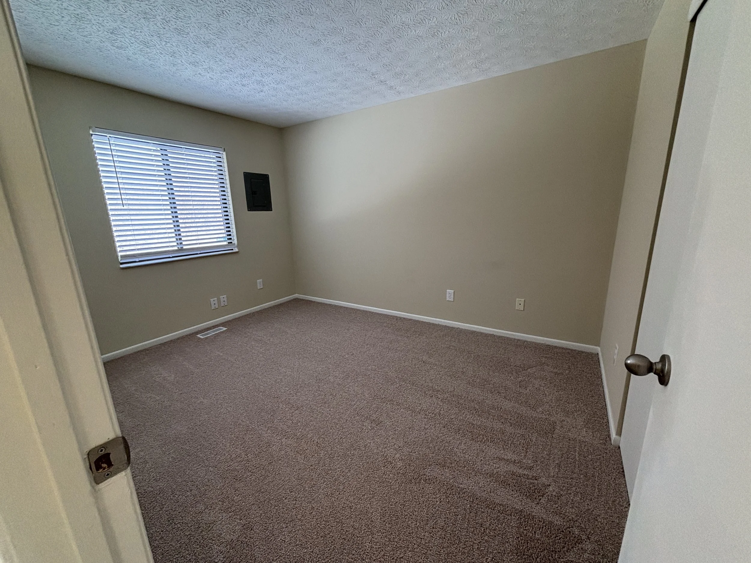 Empty room with beige walls, a window with blinds, carpet flooring, and a closed white door, with several electrical outlets on walls.
