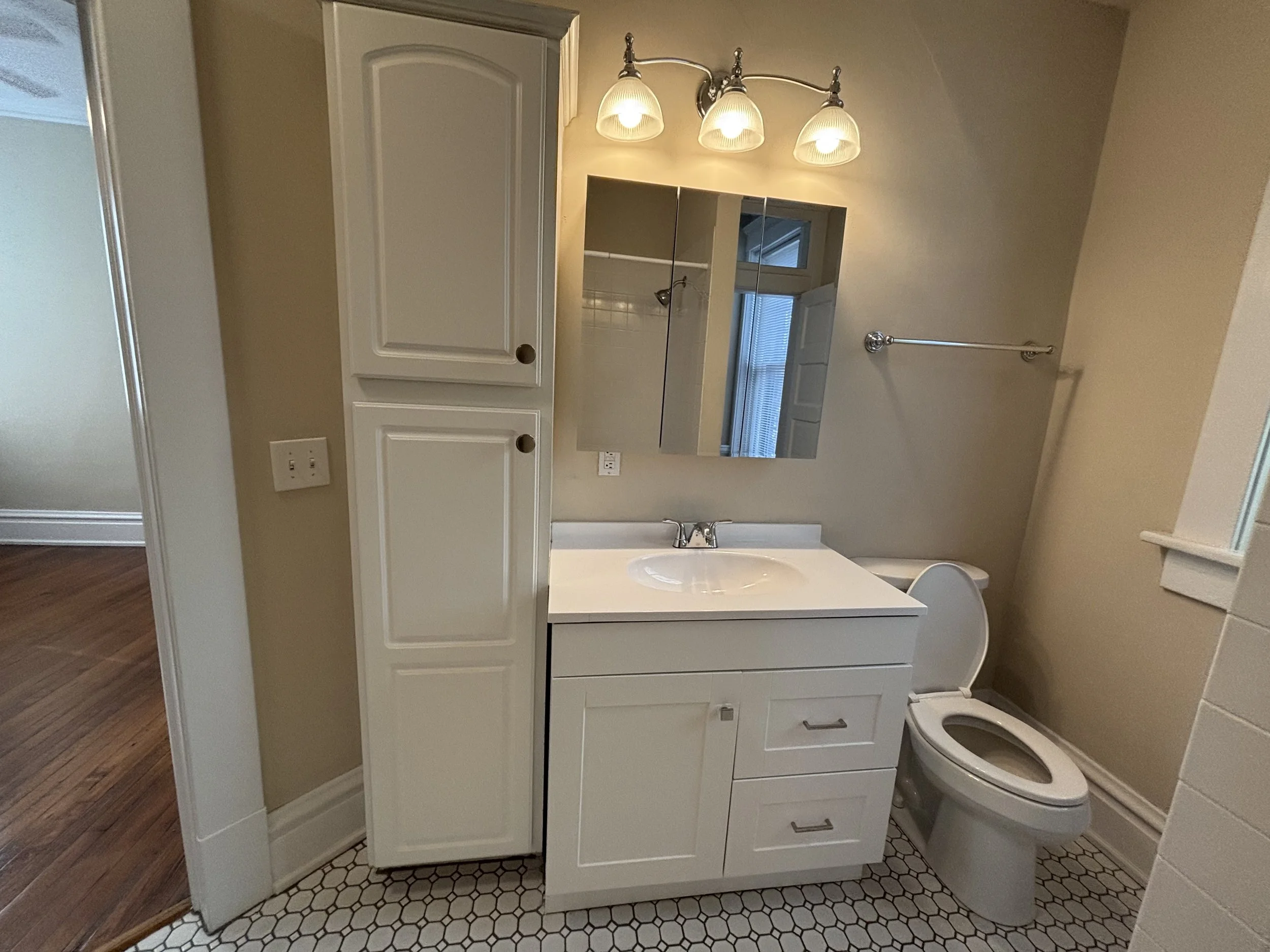 Bathroom with a white vanity, mirror, overhead light fixture, toilet, beige walls, and hexagonal tile floor.
