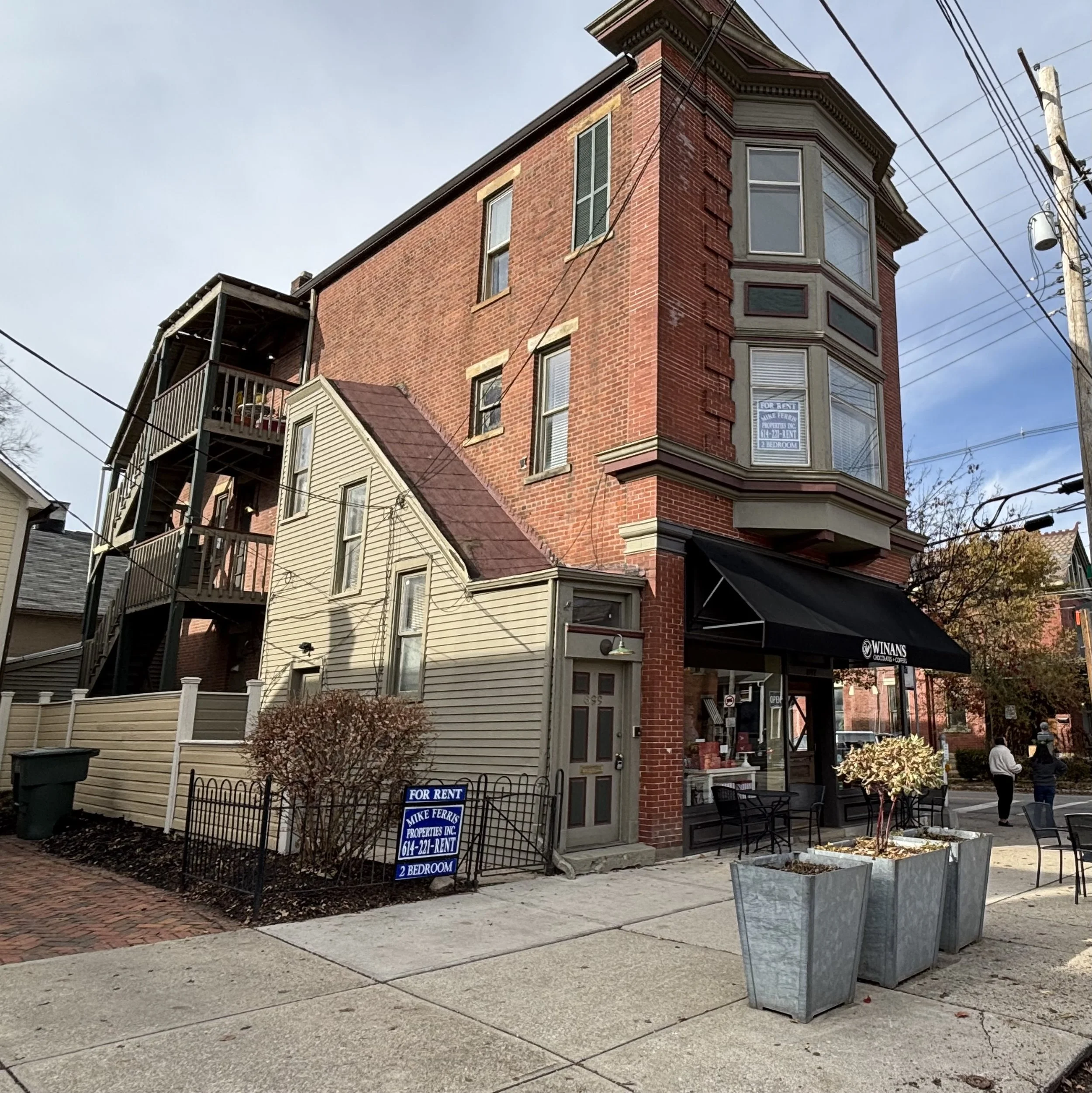 A three-story red brick building with a corner bay window and a black awning that reads 'Winans' on a city sidewalk. A blue 'For Rent' sign is in the small front yard with a bush, and two large metal planters with small trees are on the sidewalk. Peo