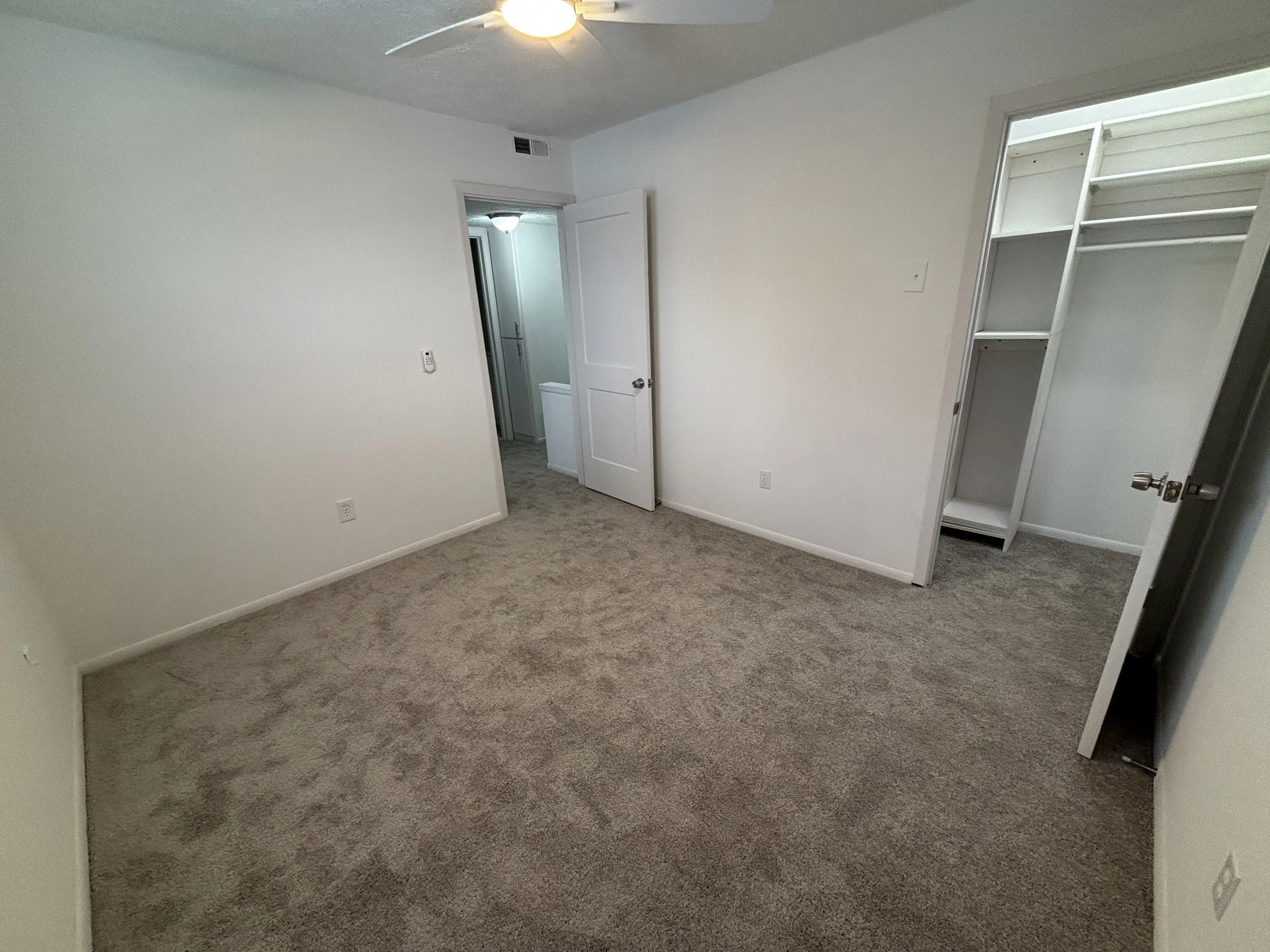 An empty bedroom with beige carpet, white walls, and a ceiling fan with a light. There are two open doors: one leading to a bathroom and the other to a closet with built-in shelves. The room appears clean and unoccupied.
