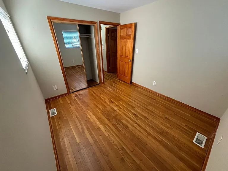 Empty bedroom with hardwood flooring, light green walls, an open closet with sliding mirror doors, a window with blinds, and a wooden door leading to another room.
