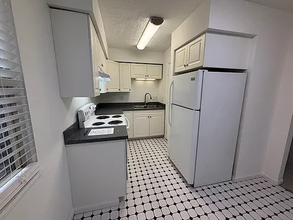 Small kitchen with white cabinets, black countertop, white refrigerator, stove, and sink in a room with patterned tile flooring and a window with blinds.