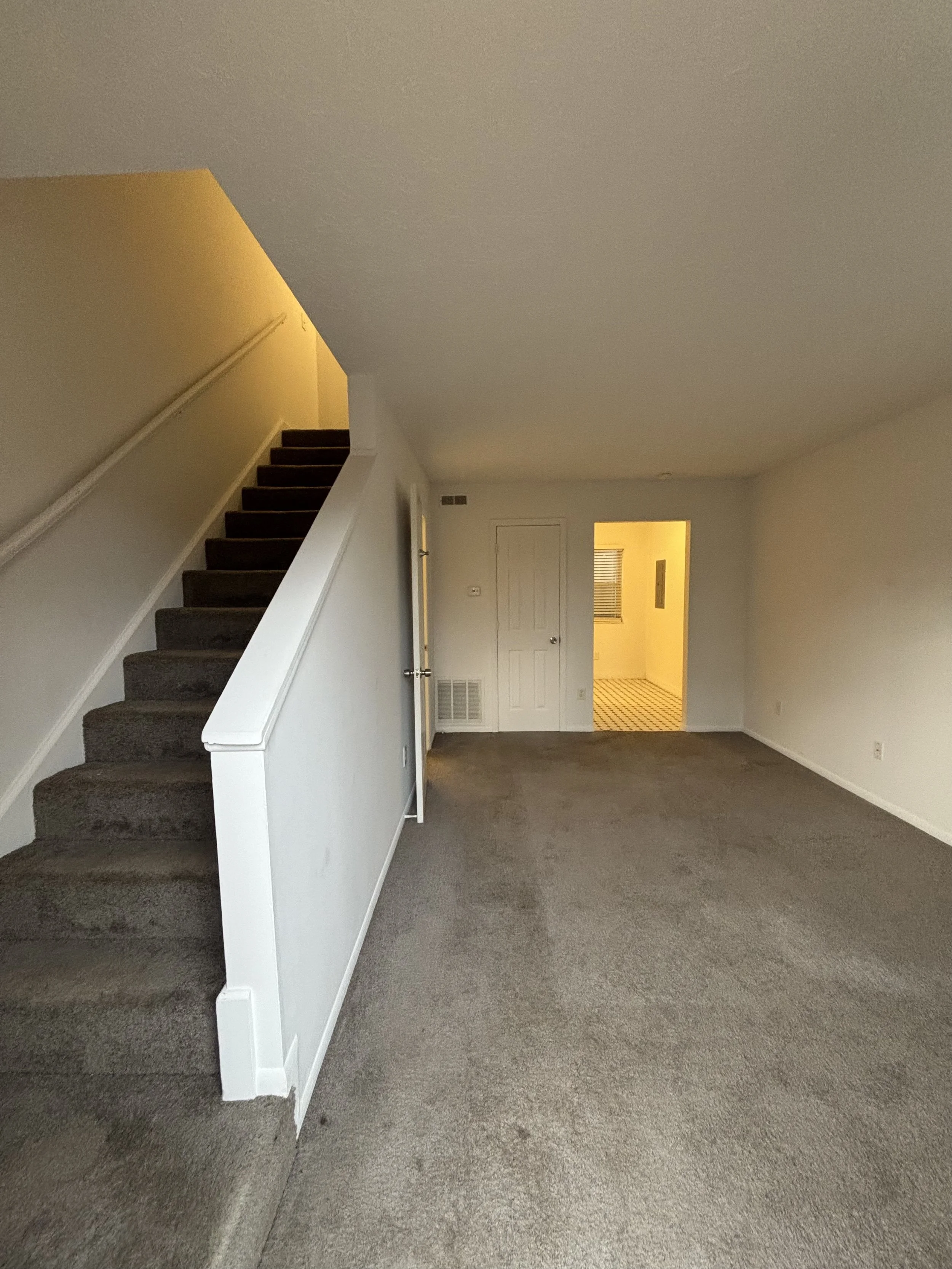 Empty living room with beige carpet, white walls, a staircase with dark carpet on the left, and an open doorway leading to a kitchen with checkered floor.