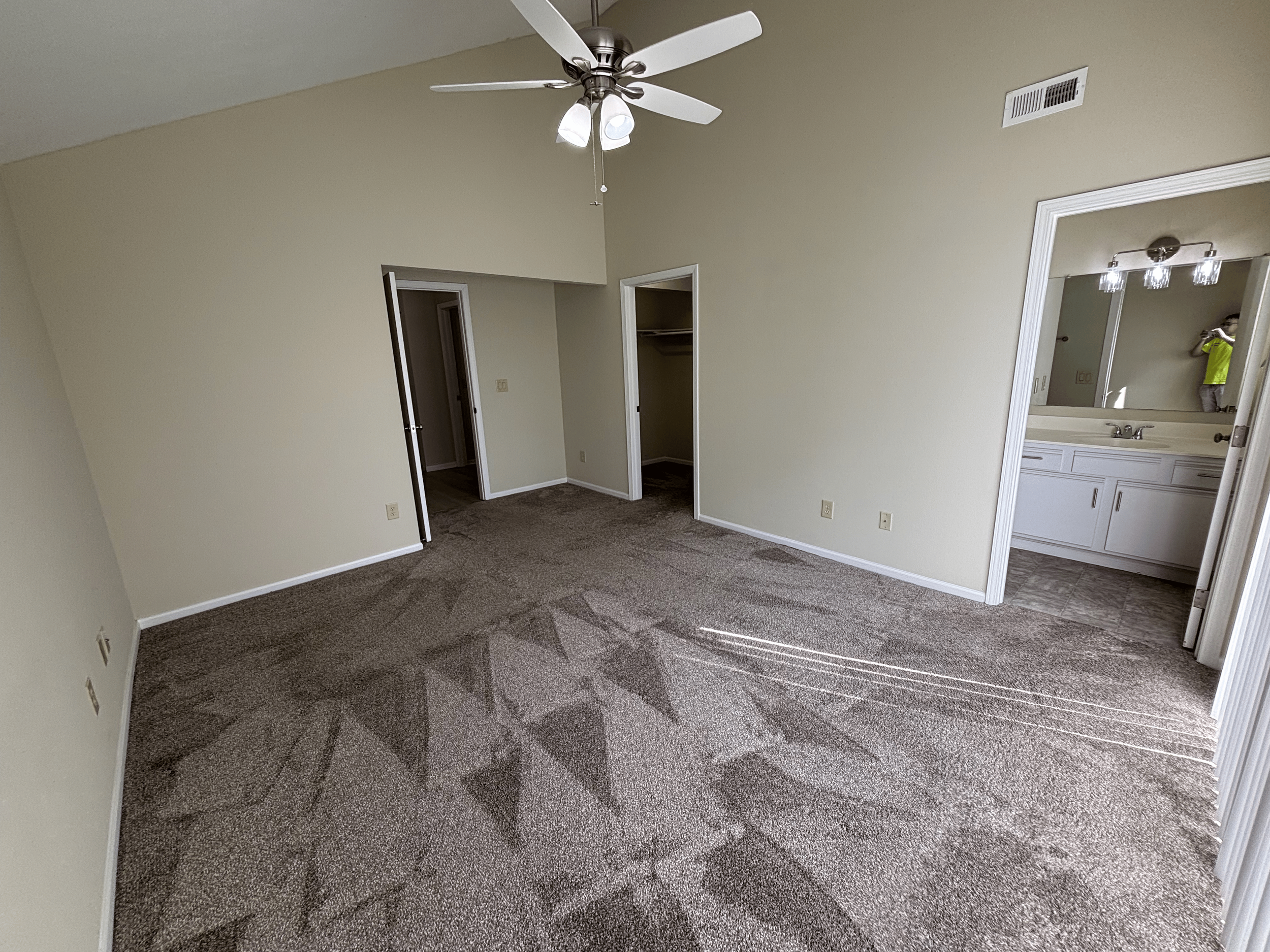 Empty bedroom with beige walls, a ceiling fan with lights, carpeted floor with vacuum cleaner lines, an open closet, and a nearby bathroom with a large mirror and white vanity.