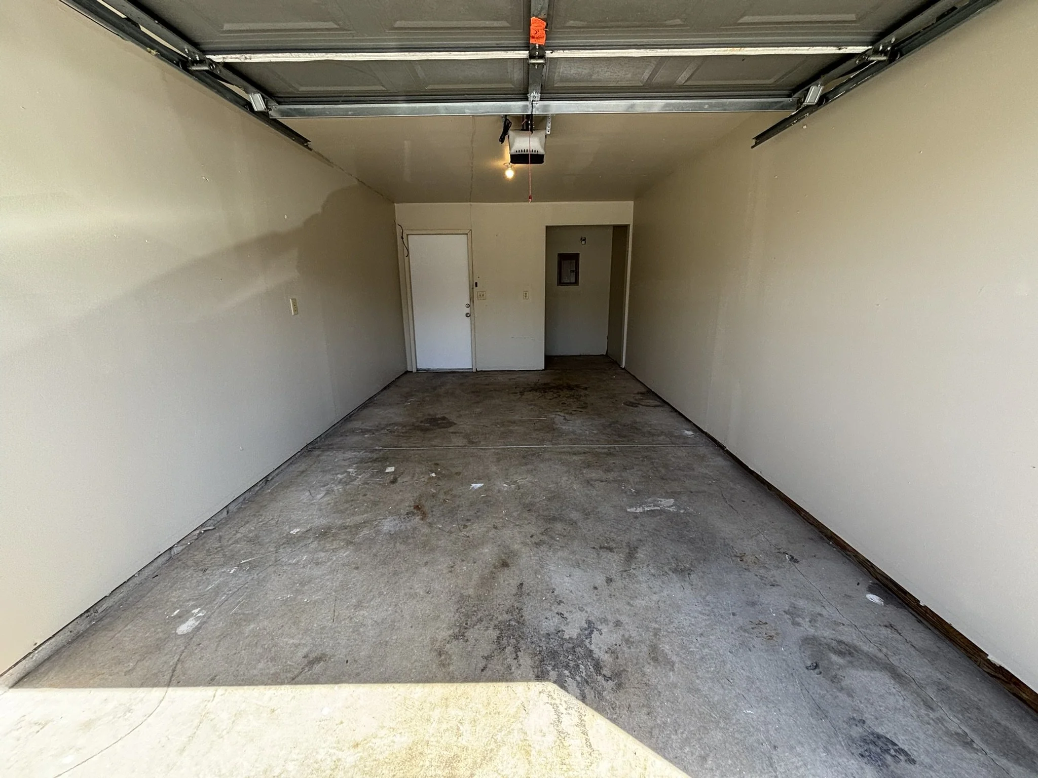 Empty garage with concrete floor, off-white walls, and a partially open garage door at the front. There is a white door at the back and a small electrical panel on the wall to the right of it.