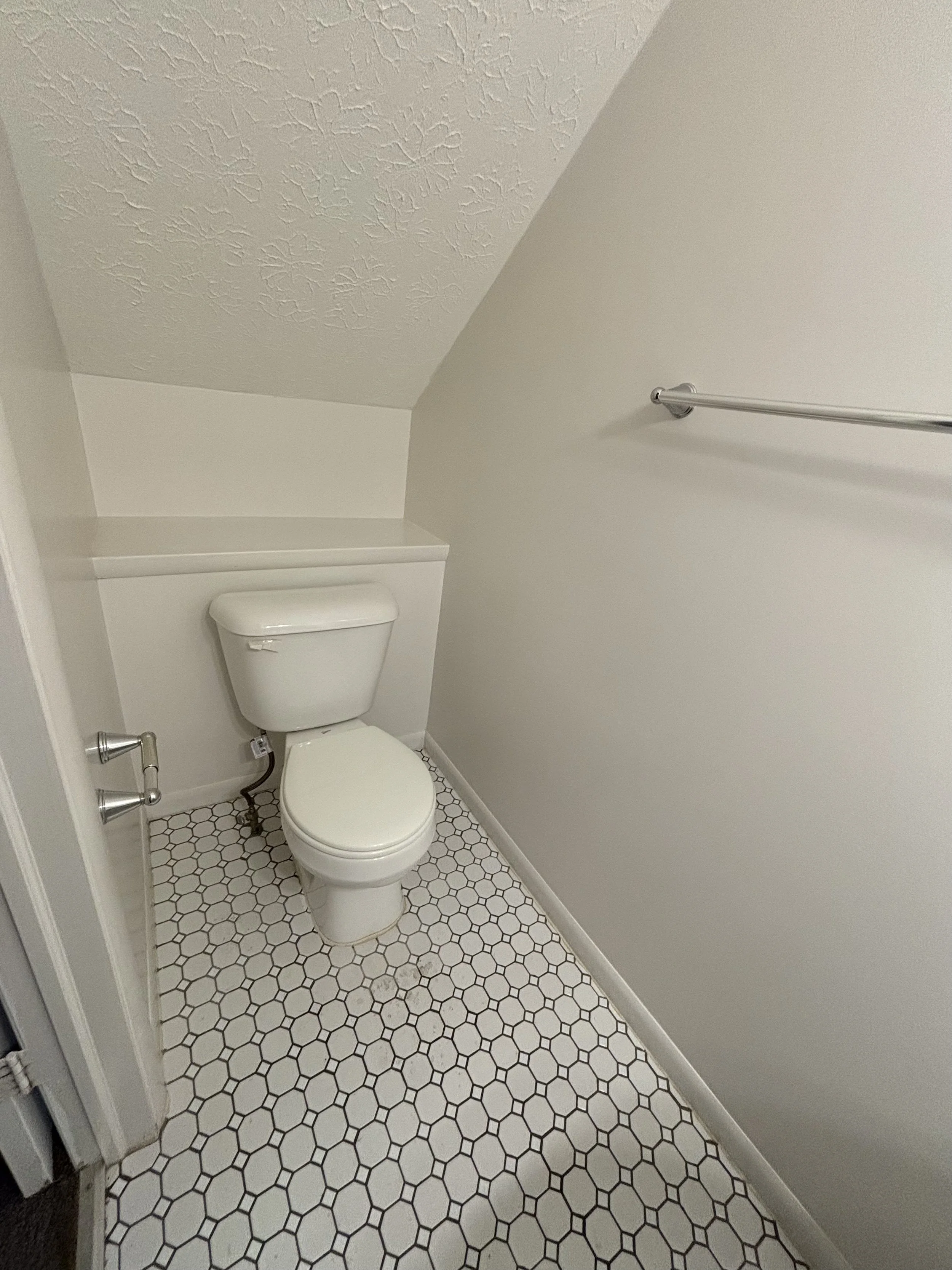 A small bathroom with white walls, a white toilet, a metal towel bar on the right wall, and a hexagon tile floor in white with black grout.